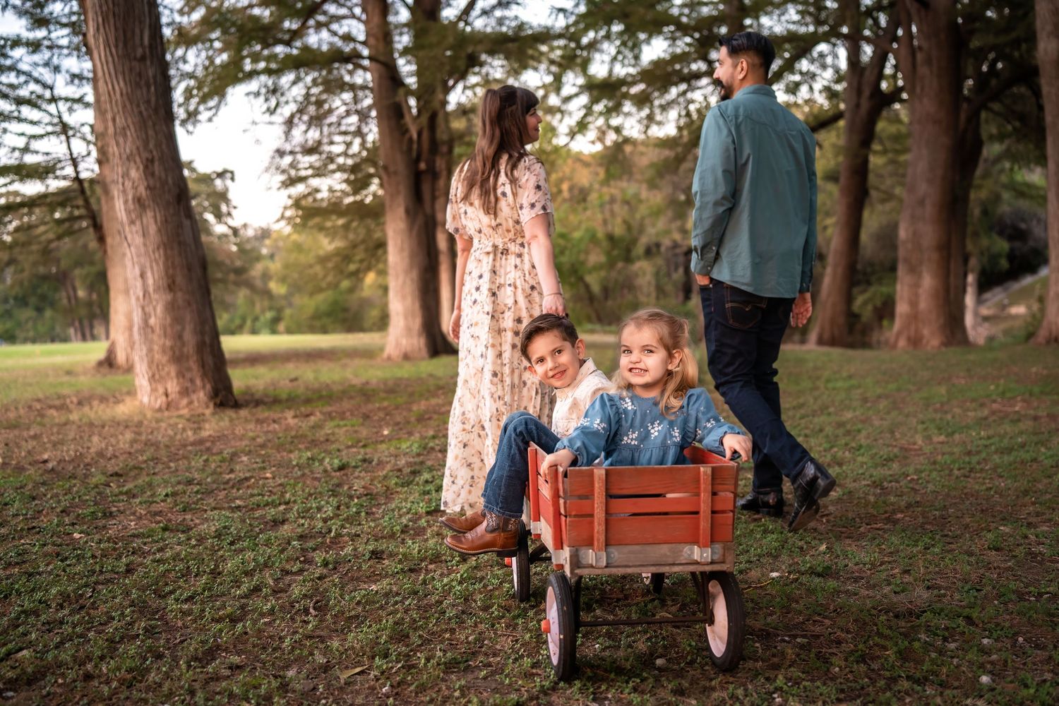 Family walking in park with two young children riding in a red wagon among tall trees during golden hour.