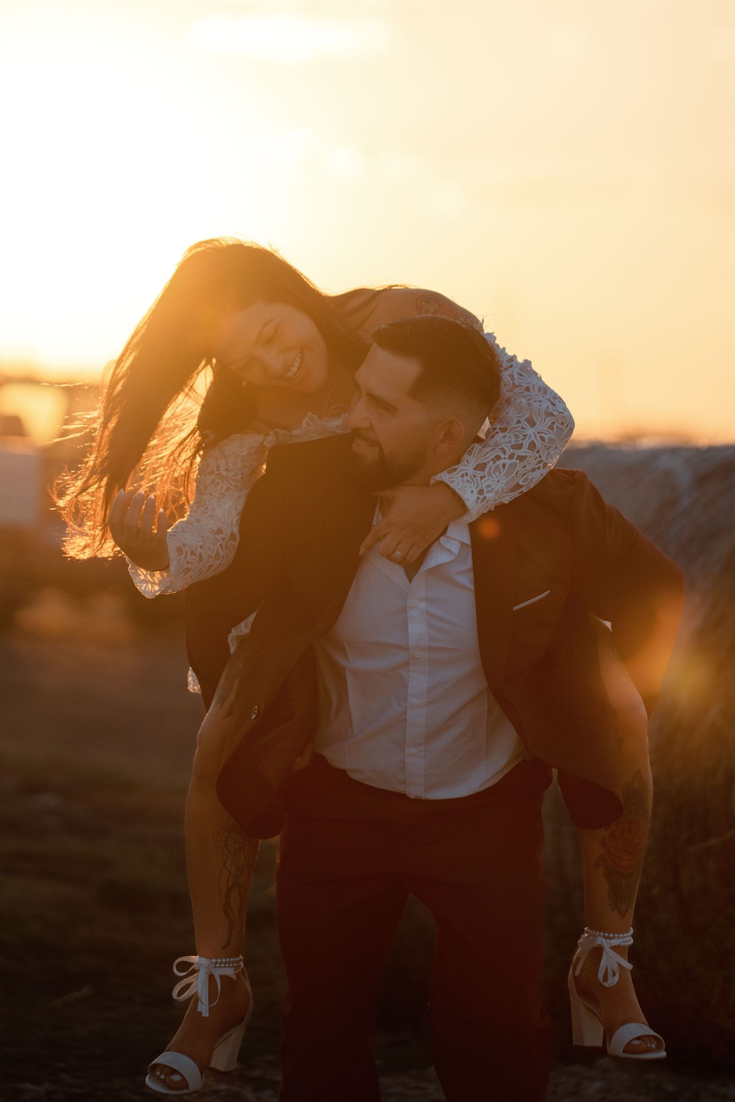 Couple sharing a romantic piggyback ride at golden sunset, woman laughing joyfully as man carries her outdoors.