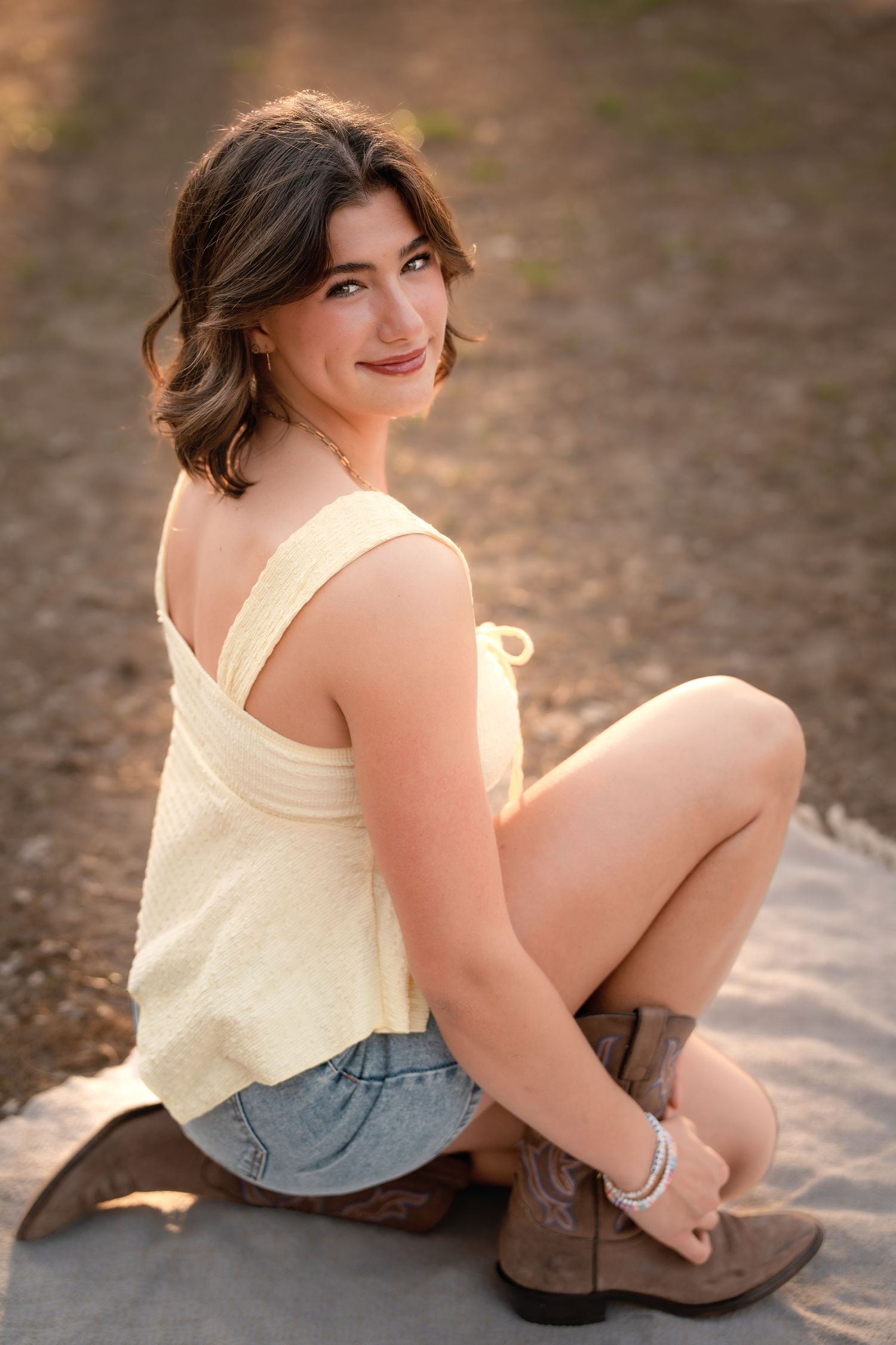 Young woman in yellow tank top, denim shorts, and cowboy boots sits outdoors, smiling over her shoulder in golden light.