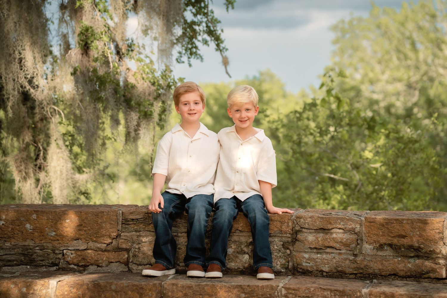 Two smiling blonde boys in white shirts and jeans sit together on a stone wall surrounded by lush green trees.
