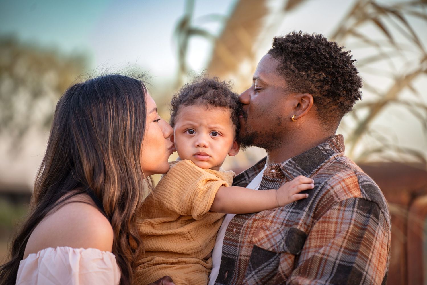 A diverse family shares tender moments together as they embrace and kiss their young child in a warm sunset setting.