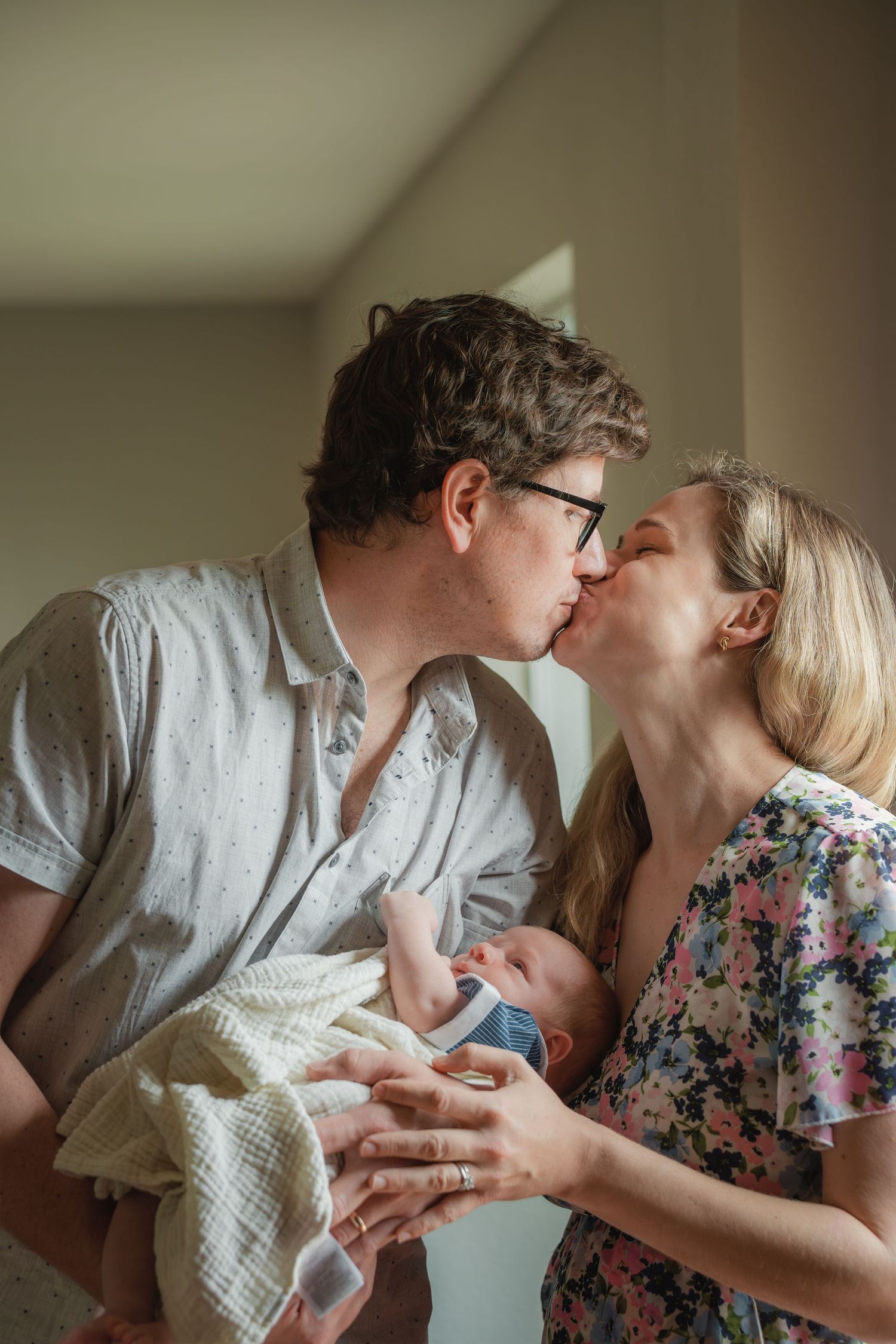 A couple shares a tender kiss while holding their newborn baby, capturing a joyful new family moment indoors.