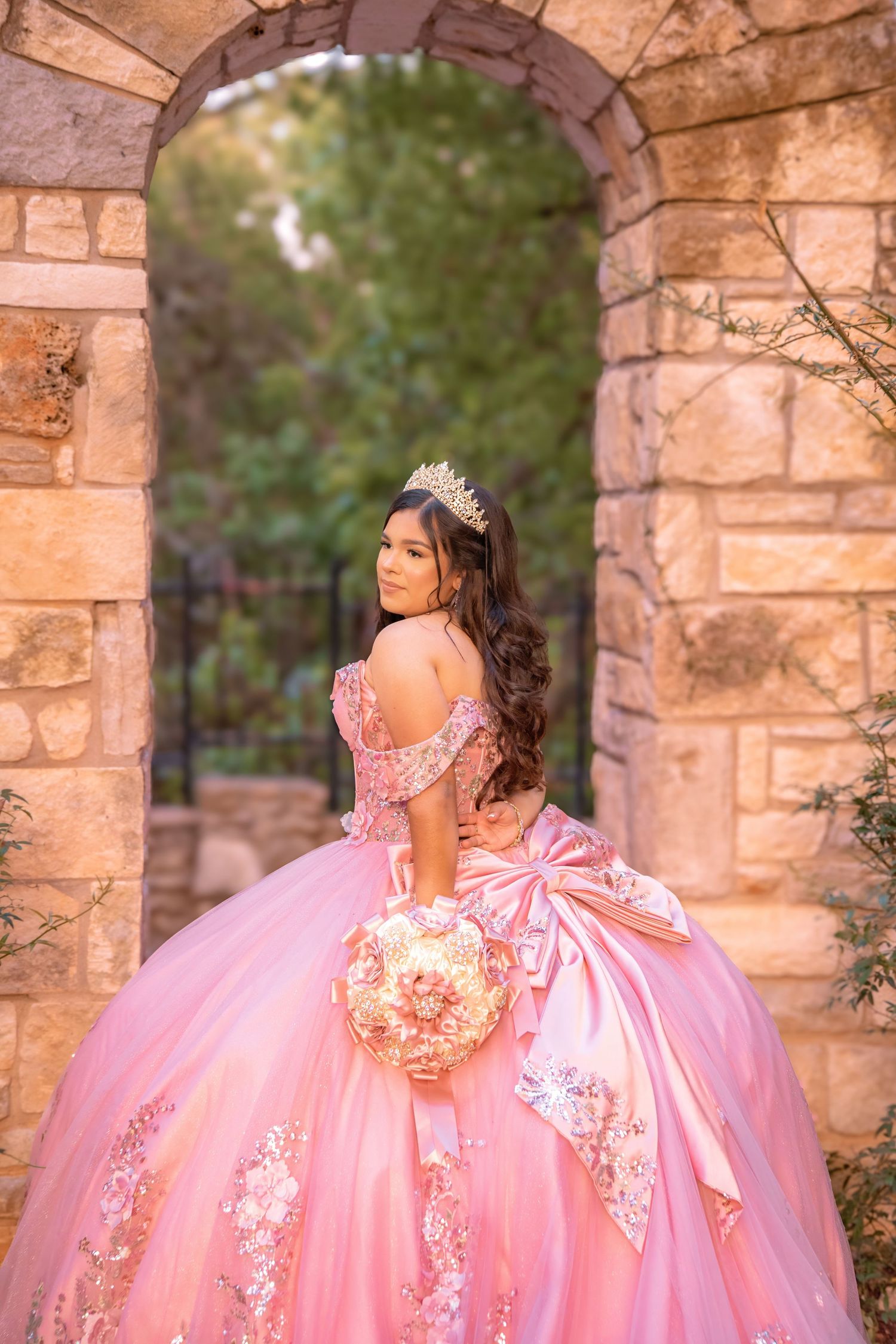 Young woman in elegant pink quinceañera ball gown with gold tiara posing under stone archway with bouquet.