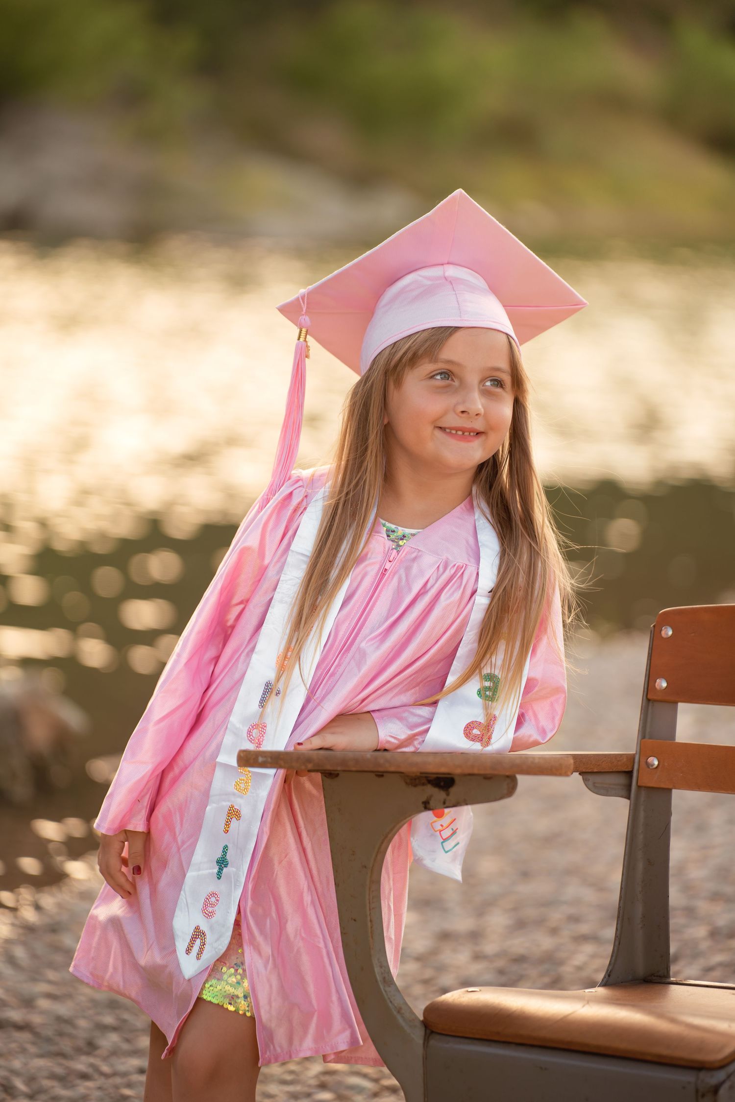 Smiling girl in pink graduation cap and gown poses proudly beside a wooden chair near a lake.