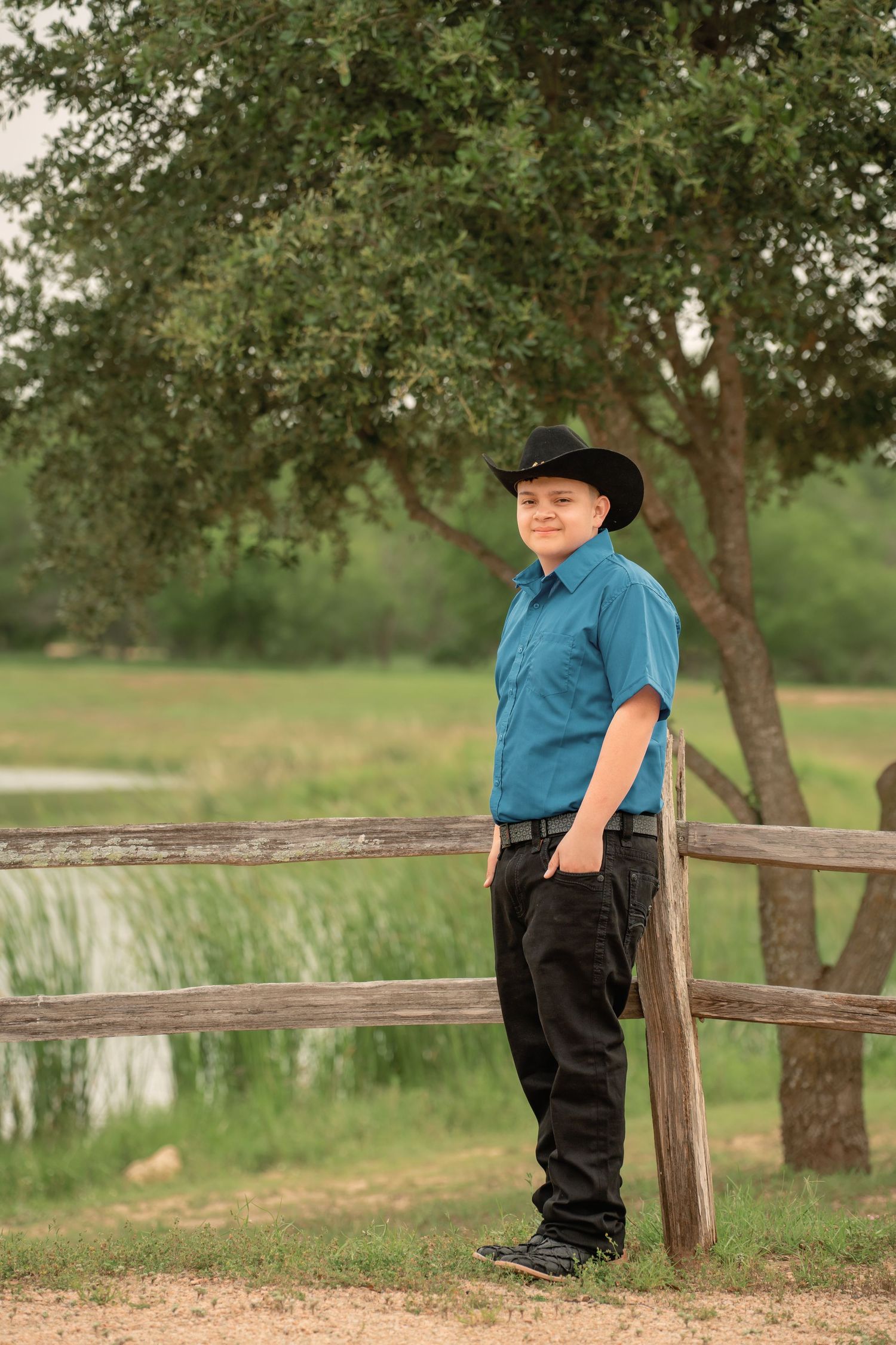 A young boy in a black cowboy hat and teal shirt leans against a wooden fence outdoors.