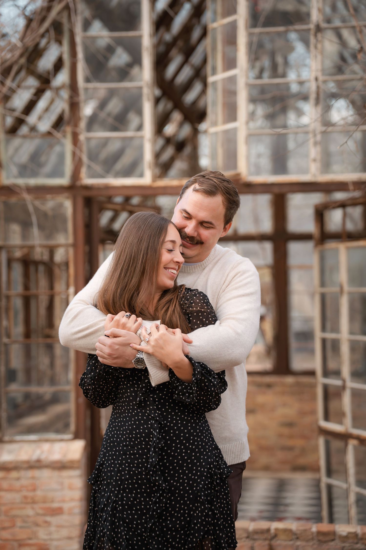A couple embraces warmly in front of a rustic greenhouse, smiling joyfully in a romantic outdoor portrait.