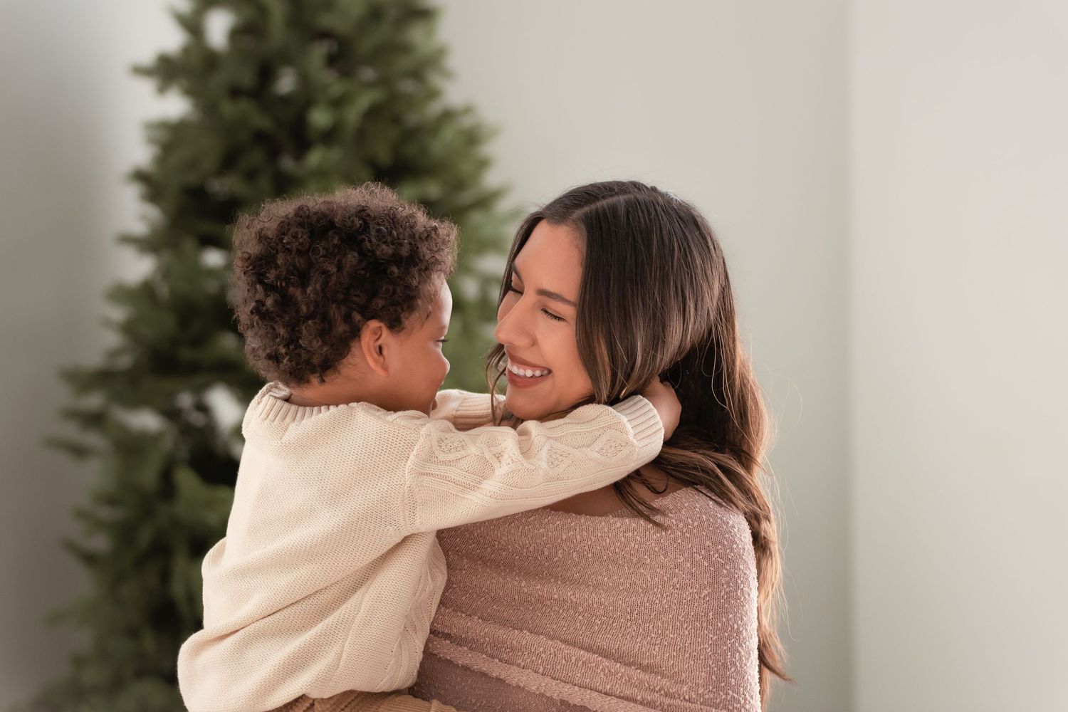 A joyful mother holds her young child in a warm embrace near a Christmas tree, both smiling and sharing a tender moment.