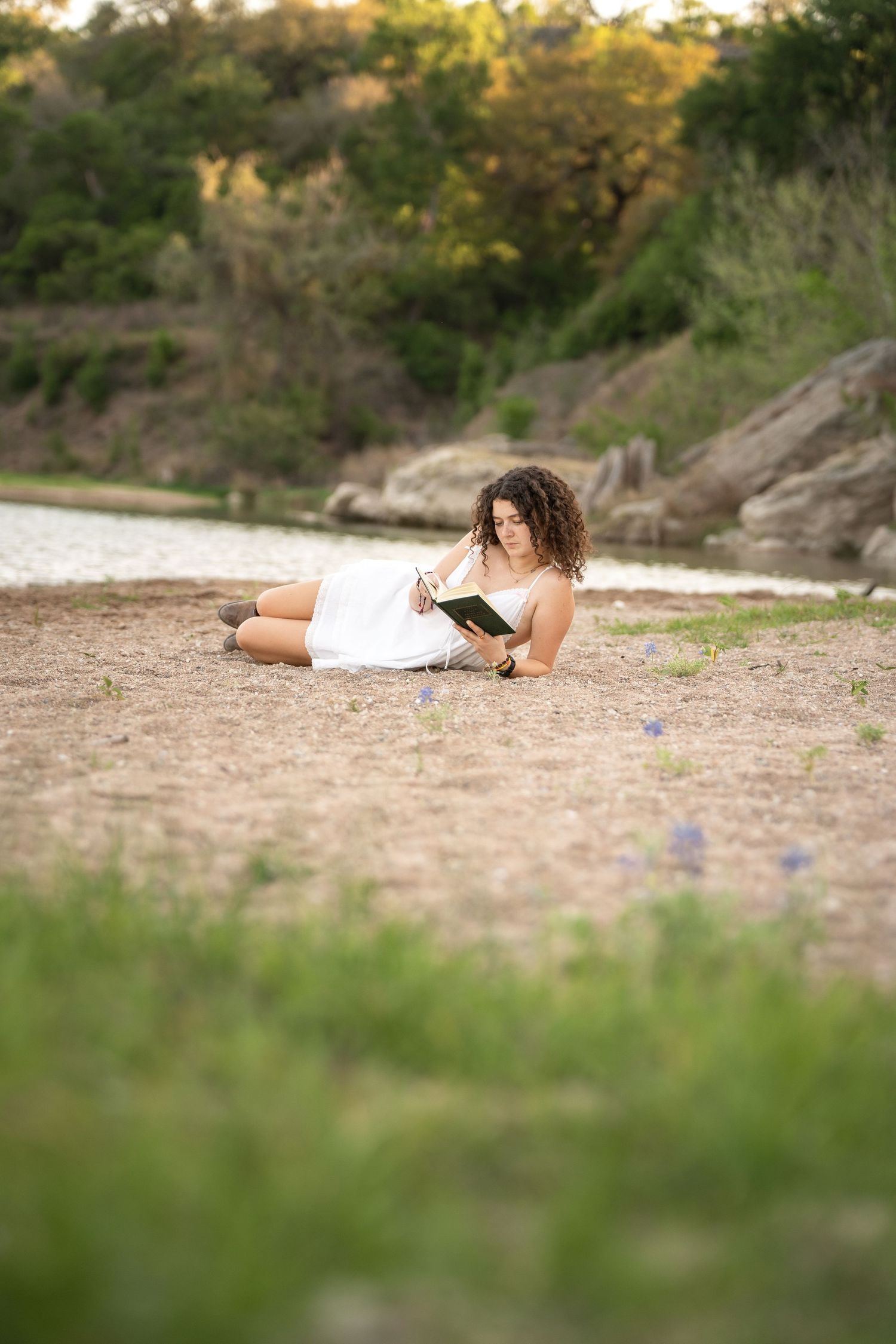 Young woman with curly hair in white dress reading a book while relaxing on sandy ground near a river.