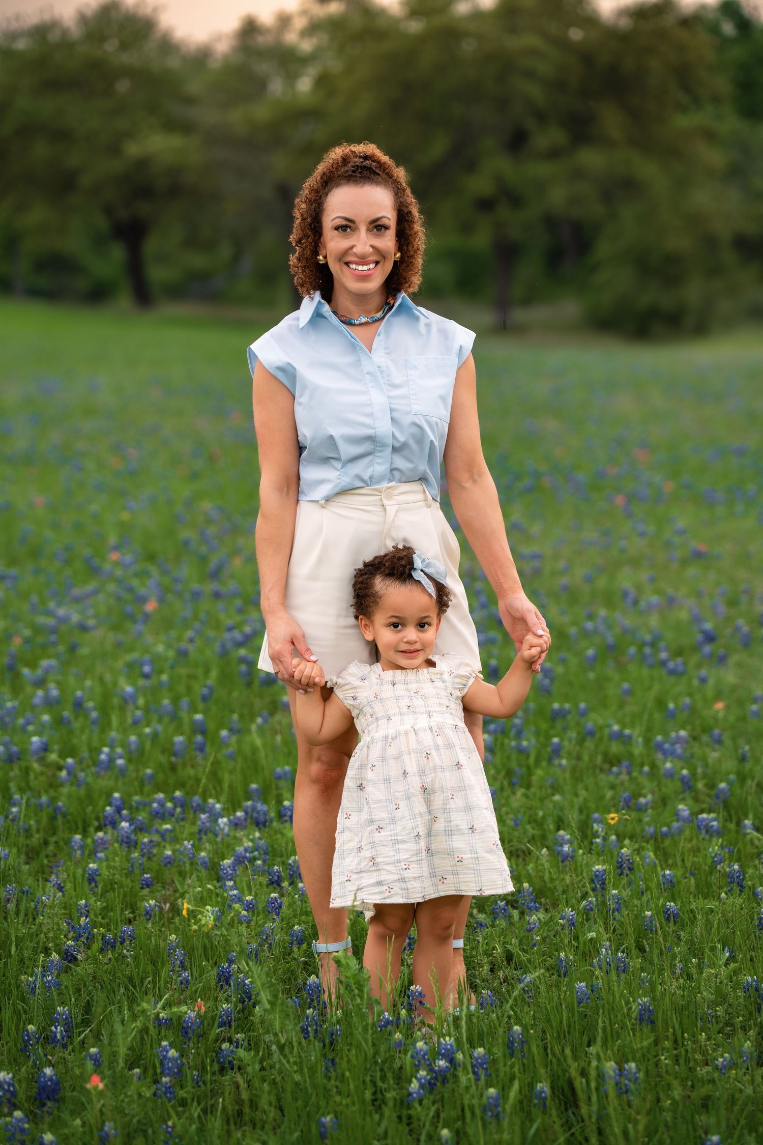 A parent and child walk together through a vibrant field of blooming bluebonnets at sunset.