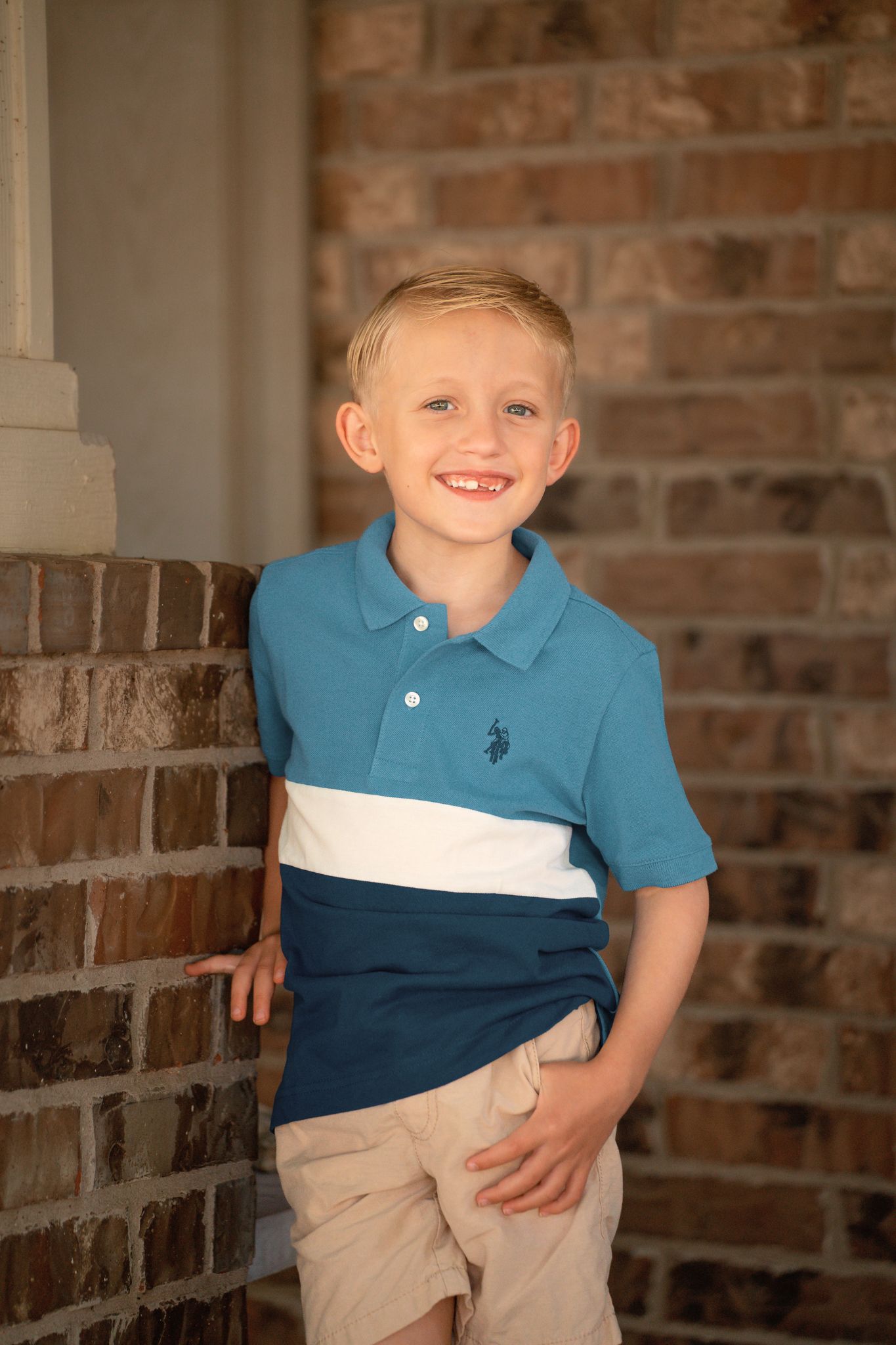 Smiling blonde boy in blue polo shirt and khaki shorts leaning against brick wall outdoors.