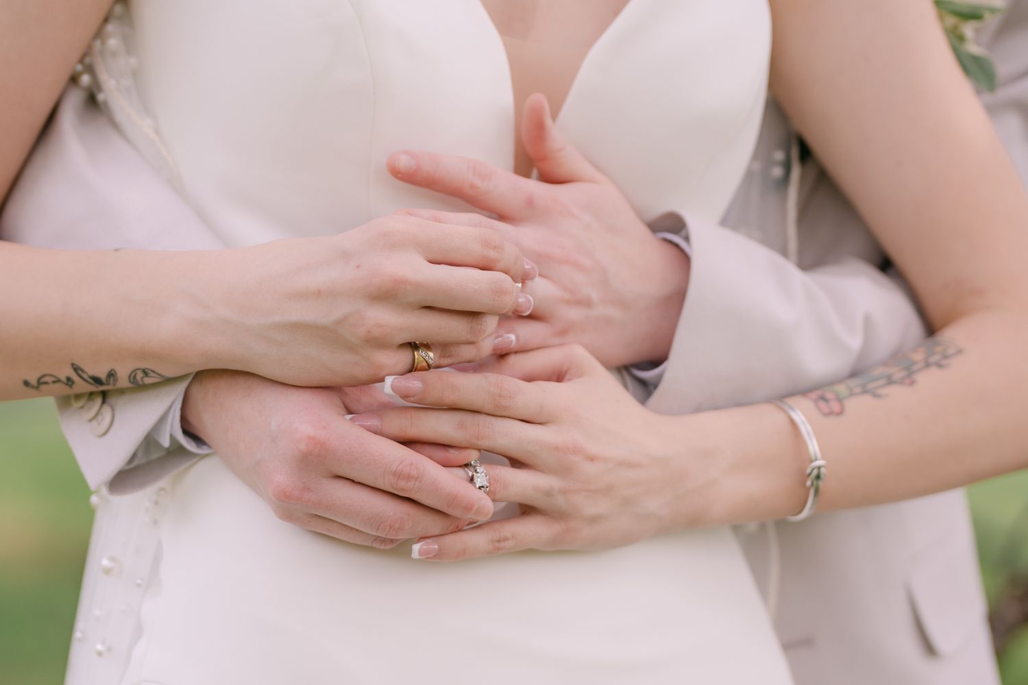 Close up of hands with gold rings and bracelets embracing against a white dress and soft green background.