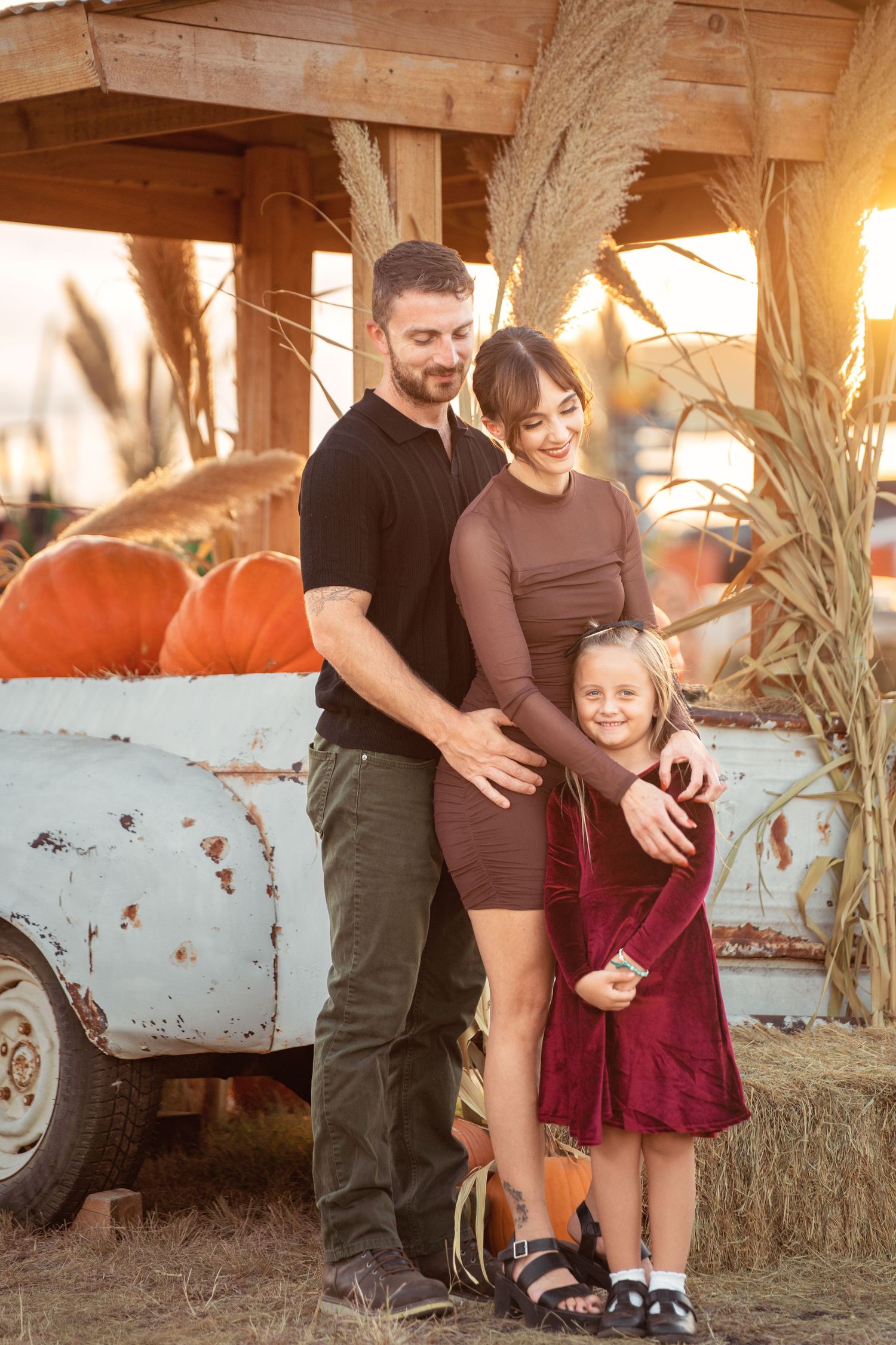 Family of three posing at a pumpkin patch in autumn, with a vintage truck, corn stalks, and warm golden sunset light.