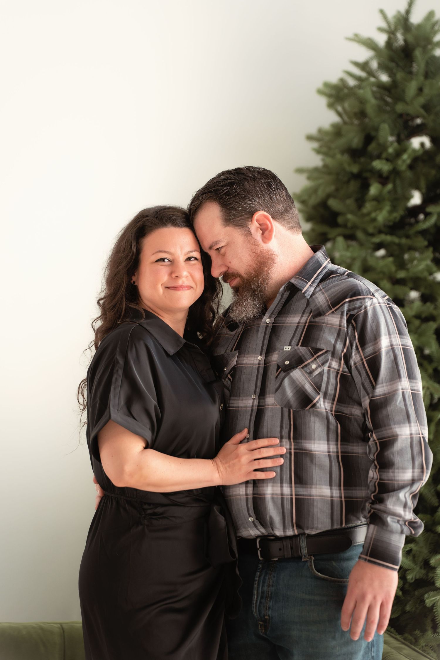 Couple sharing a tender moment by a Christmas tree, woman in black jumpsuit smiling at camera with man in plaid.