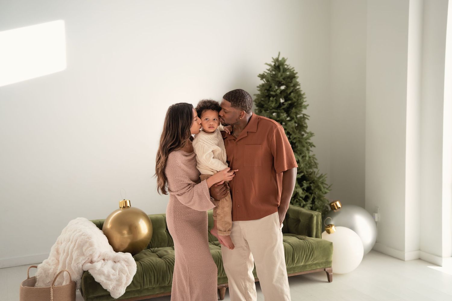 Family with baby posing near decorated Christmas tree in bright white living room with green sofa.