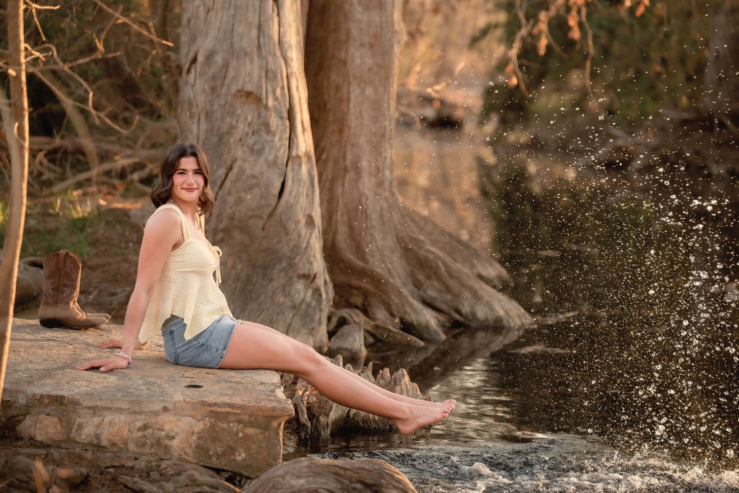 Young woman in denim shorts sitting on rocks by a flowing river surrounded by trees in golden sunlight.