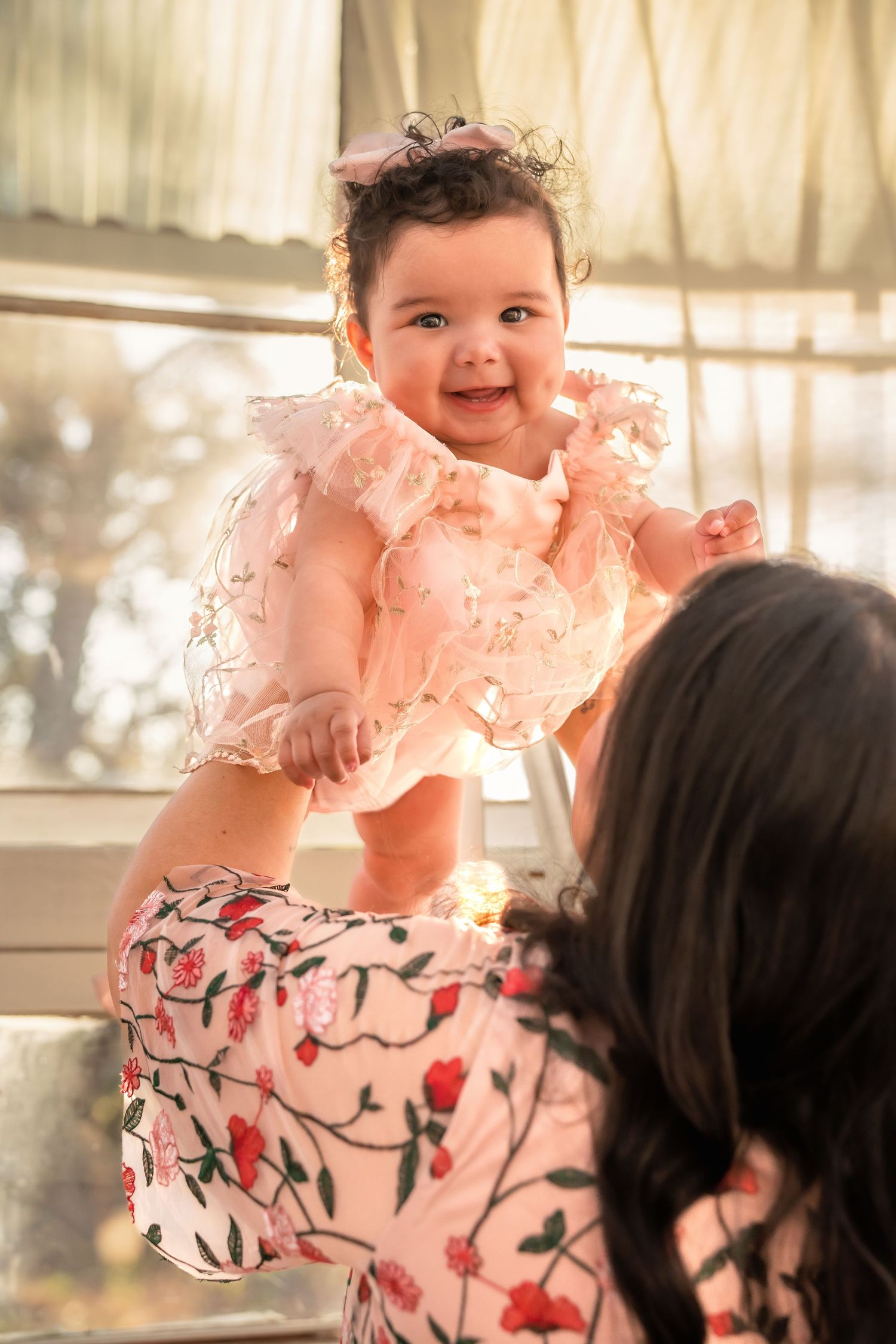 A joyful baby in a pink ruffled dress being held up by a woman in a floral outfit near a bright window.