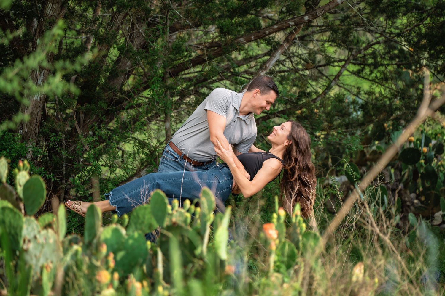 Couple sharing a romantic dip pose surrounded by lush green trees and cactus plants outdoors.
