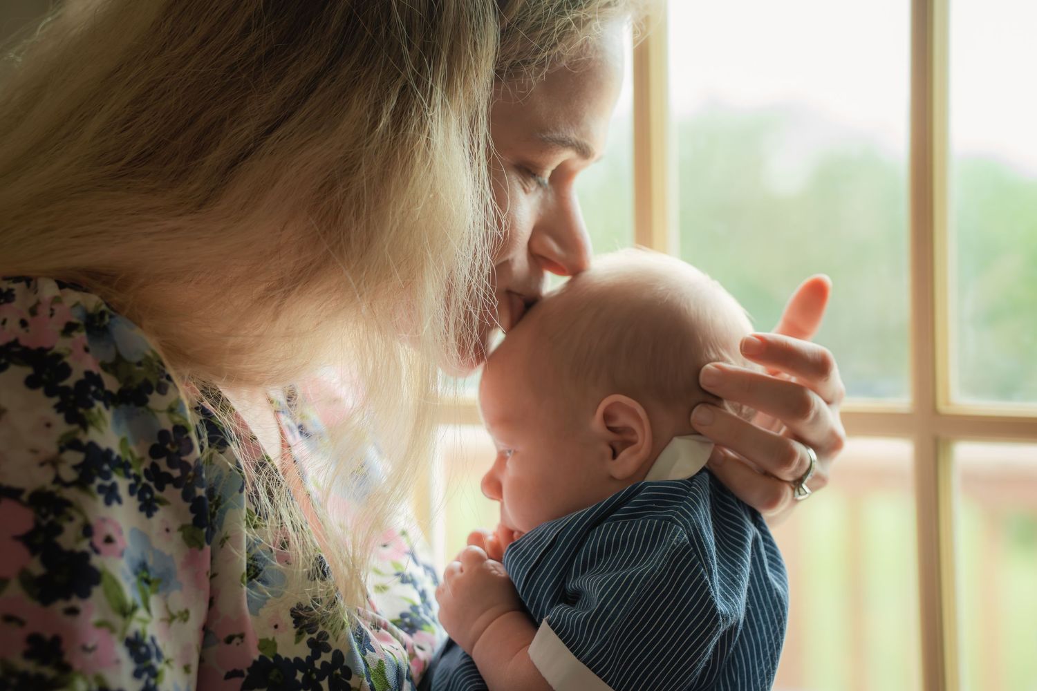 A tender moment as a blonde mother gently nuzzles her newborn baby by a sunlit window.