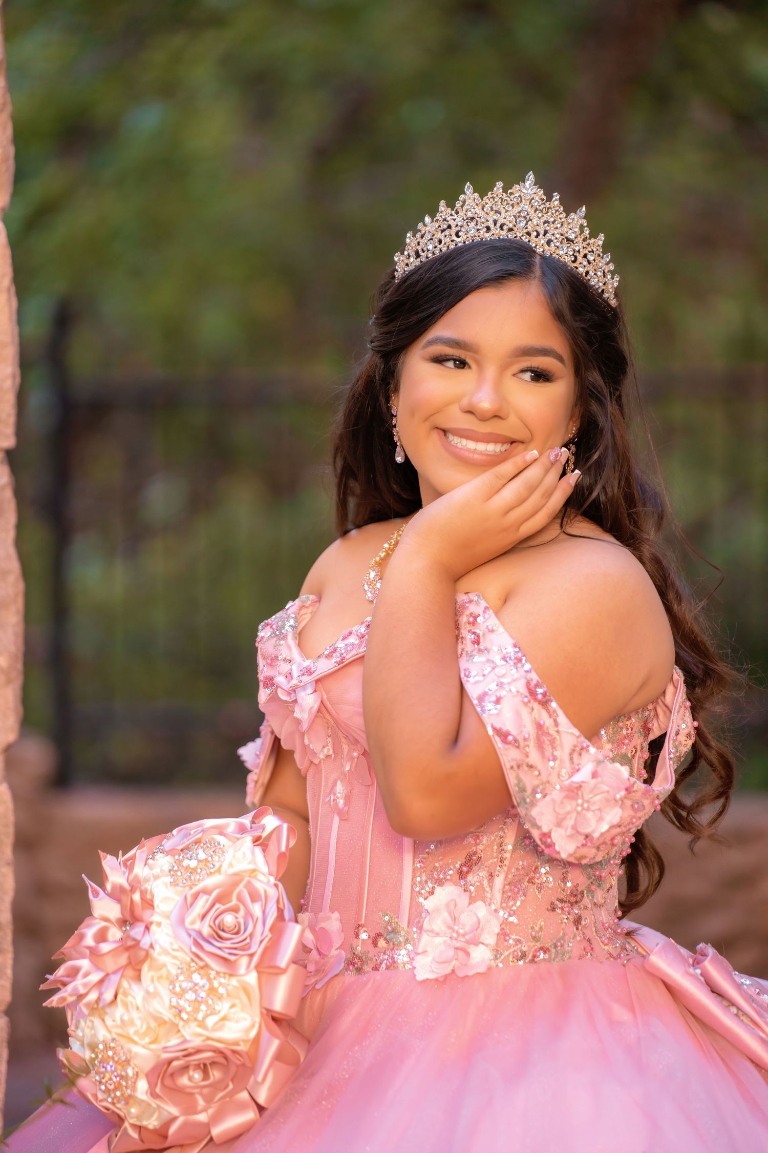 Smiling young woman in pink quinceañera gown with gold tiara holding floral bouquet, posing elegantly outdoors.