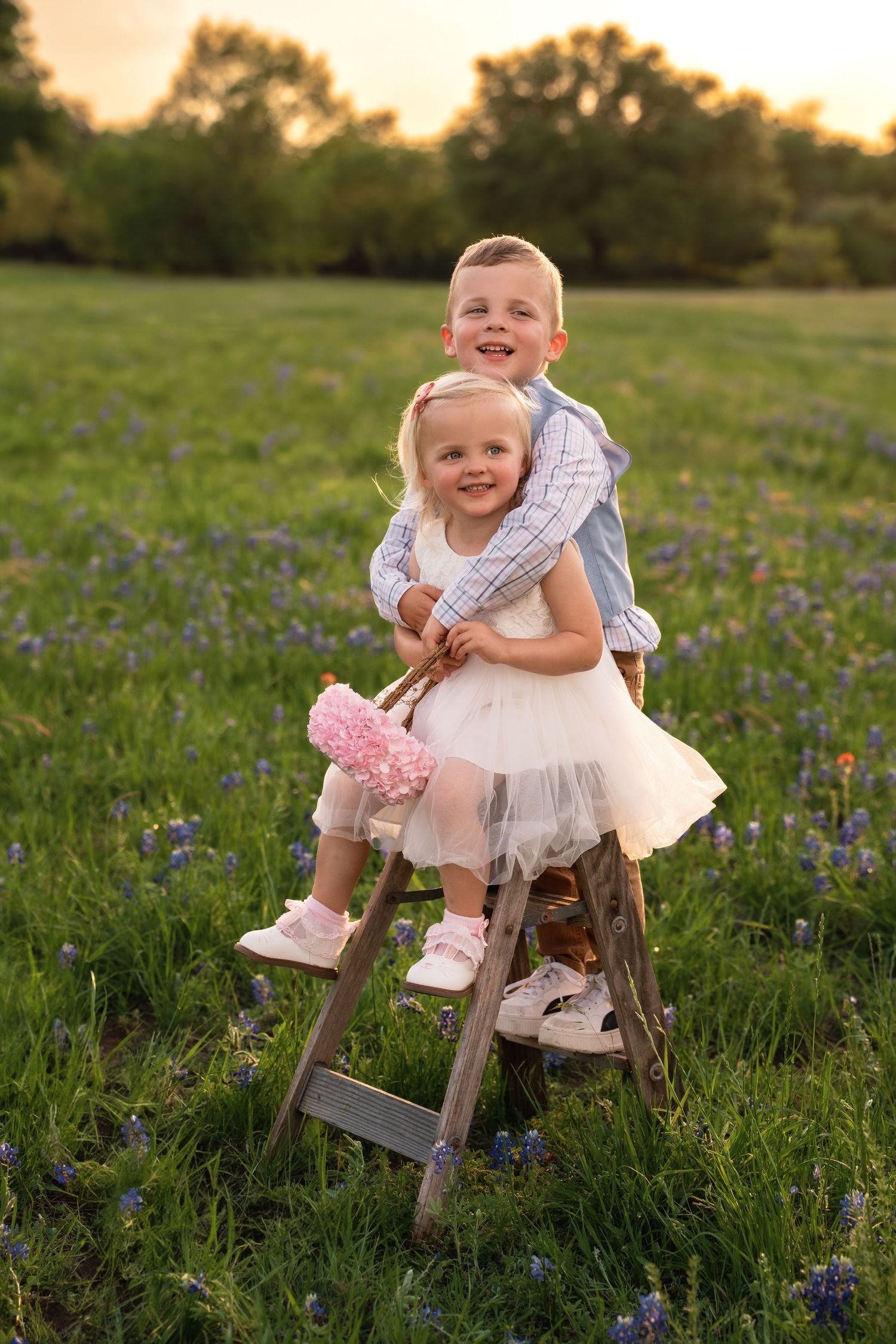 A baby in a white dress sits on a rustic wooden stump in a field of bluebonnets during golden hour.
