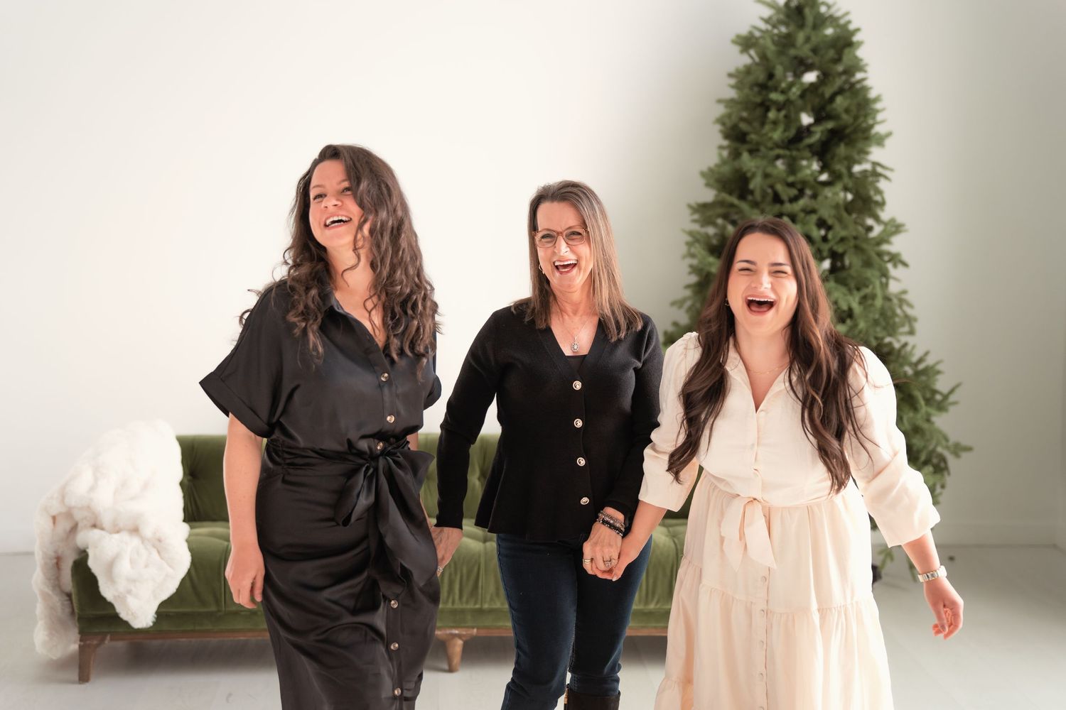 Three women laughing and holding hands in front of a Christmas tree in a festive holiday photo.