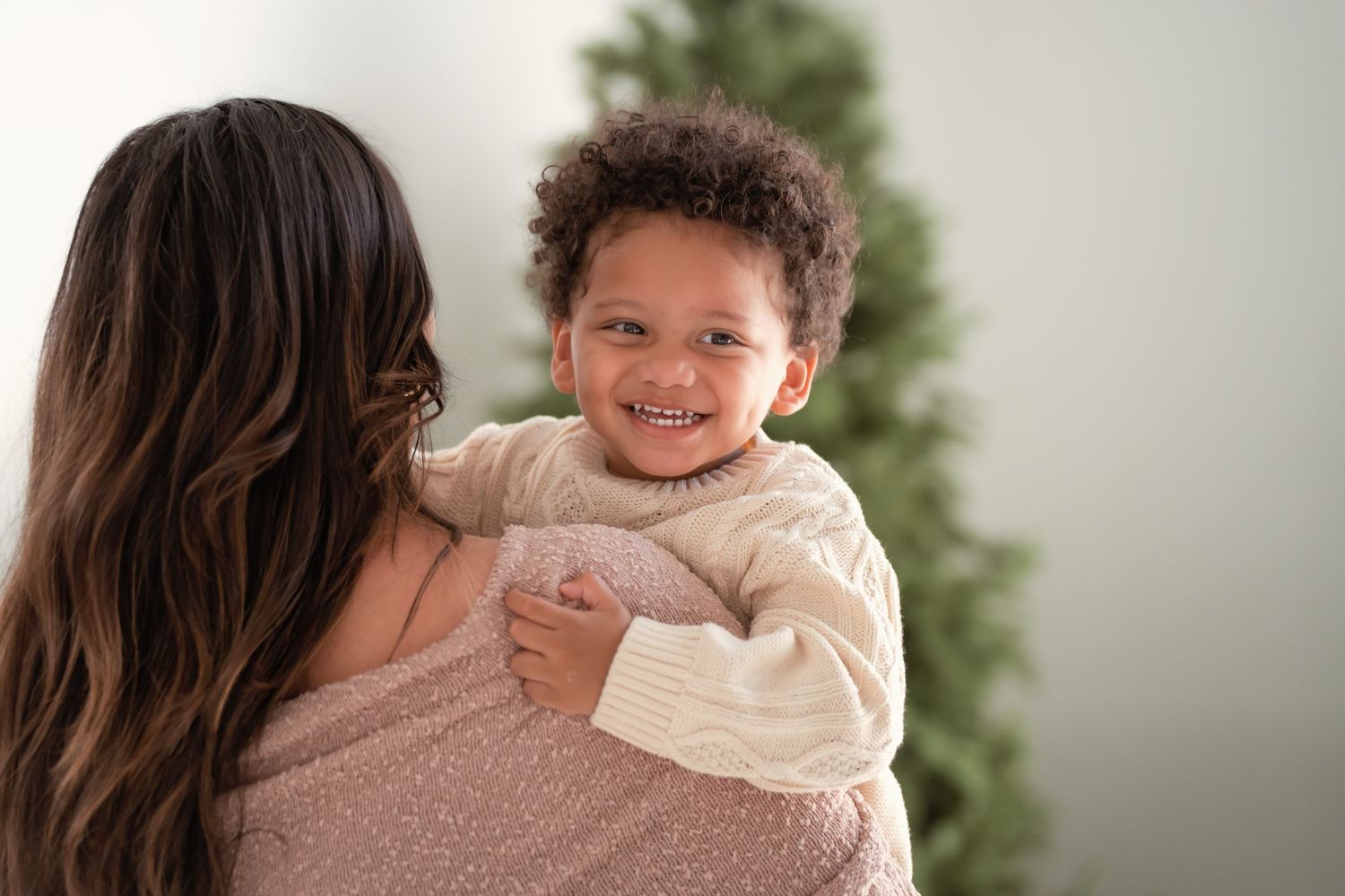 A smiling toddler in a cream sweater being held by a woman with long dark hair near a Christmas tree.