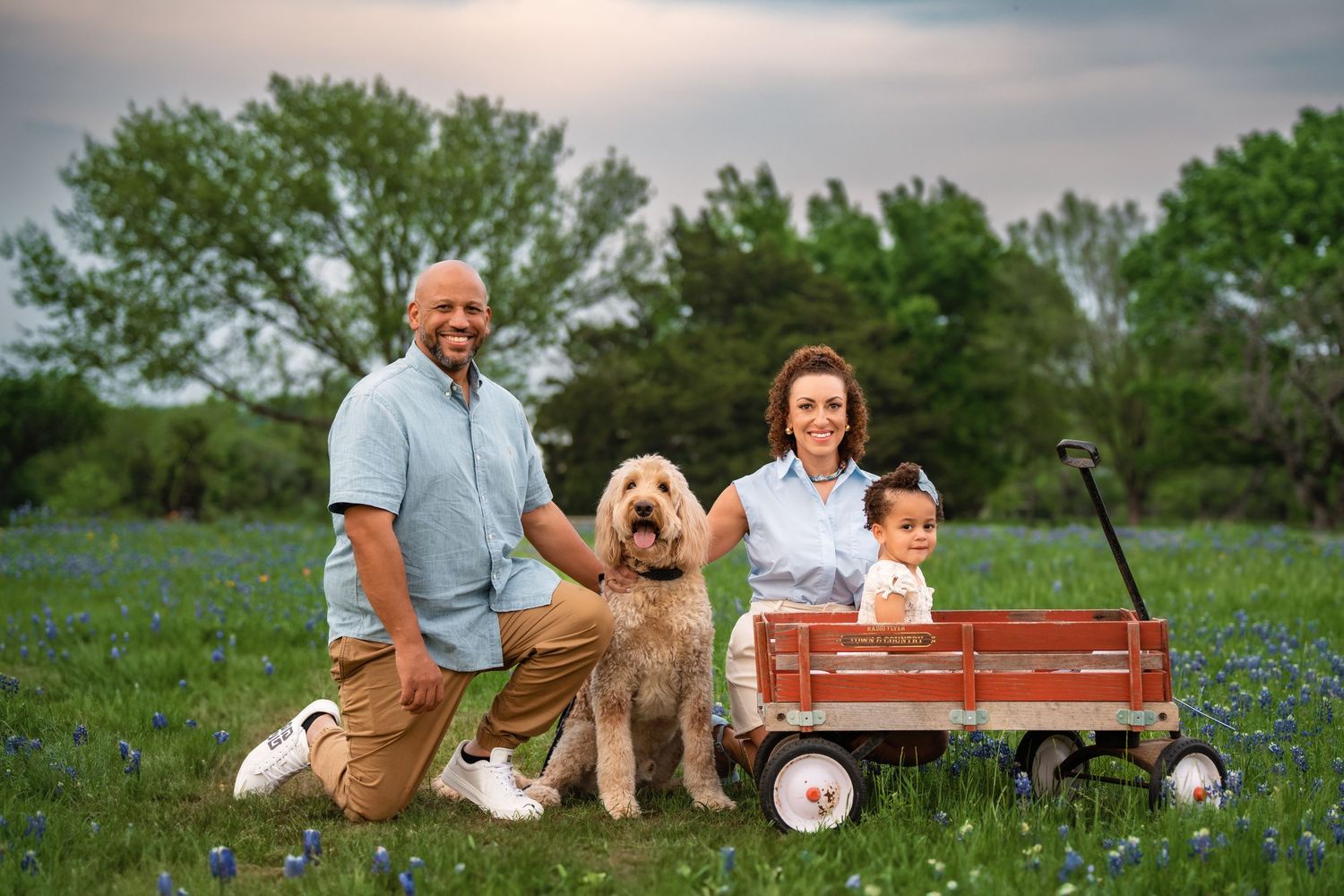 A family and their goldendoodle dog pose together with a red wagon in a field of blue wildflowers at sunset.