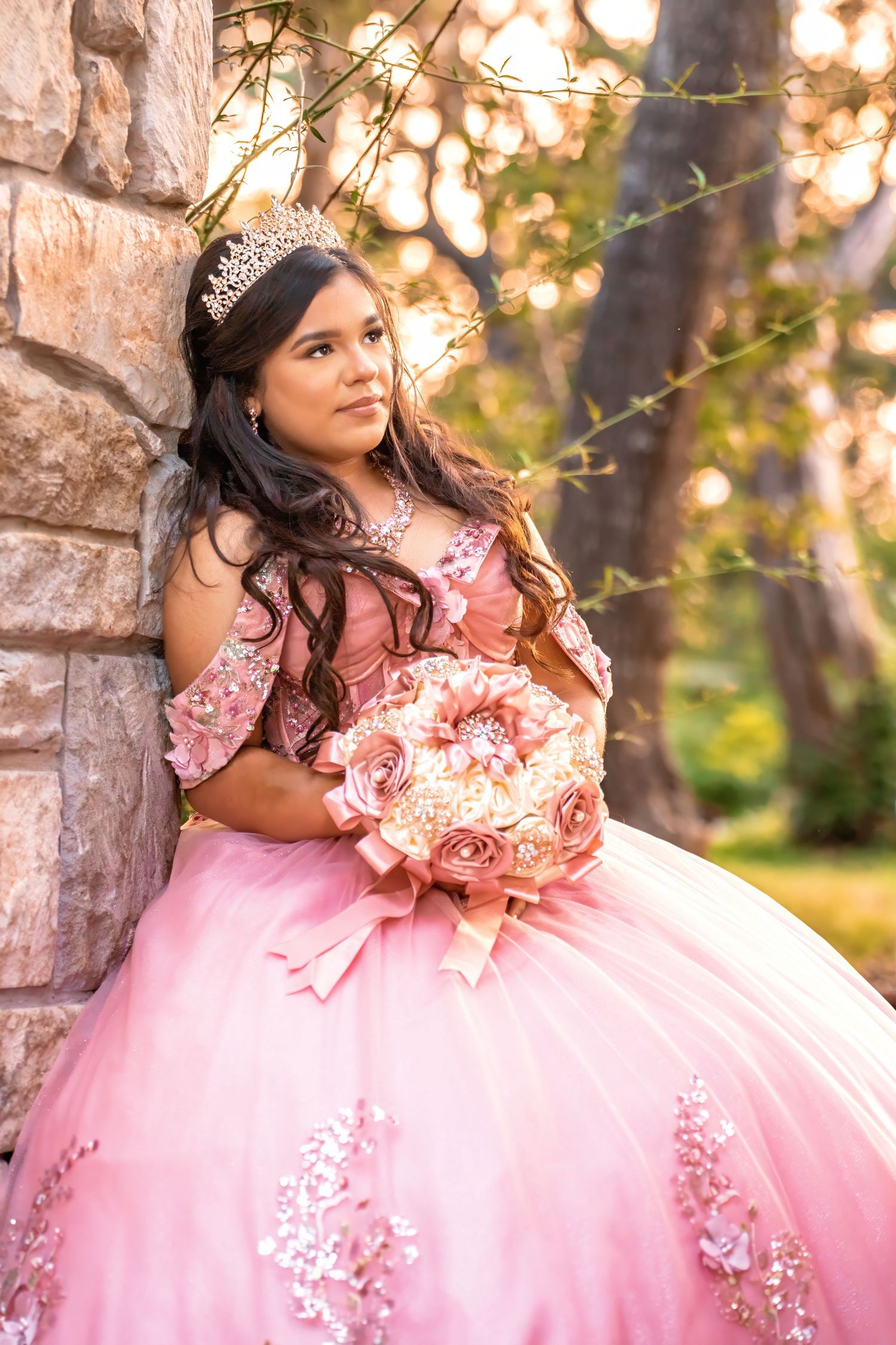 Young Latina in pink quinceañera ball gown with tiara holds floral bouquet outdoors by stone wall.