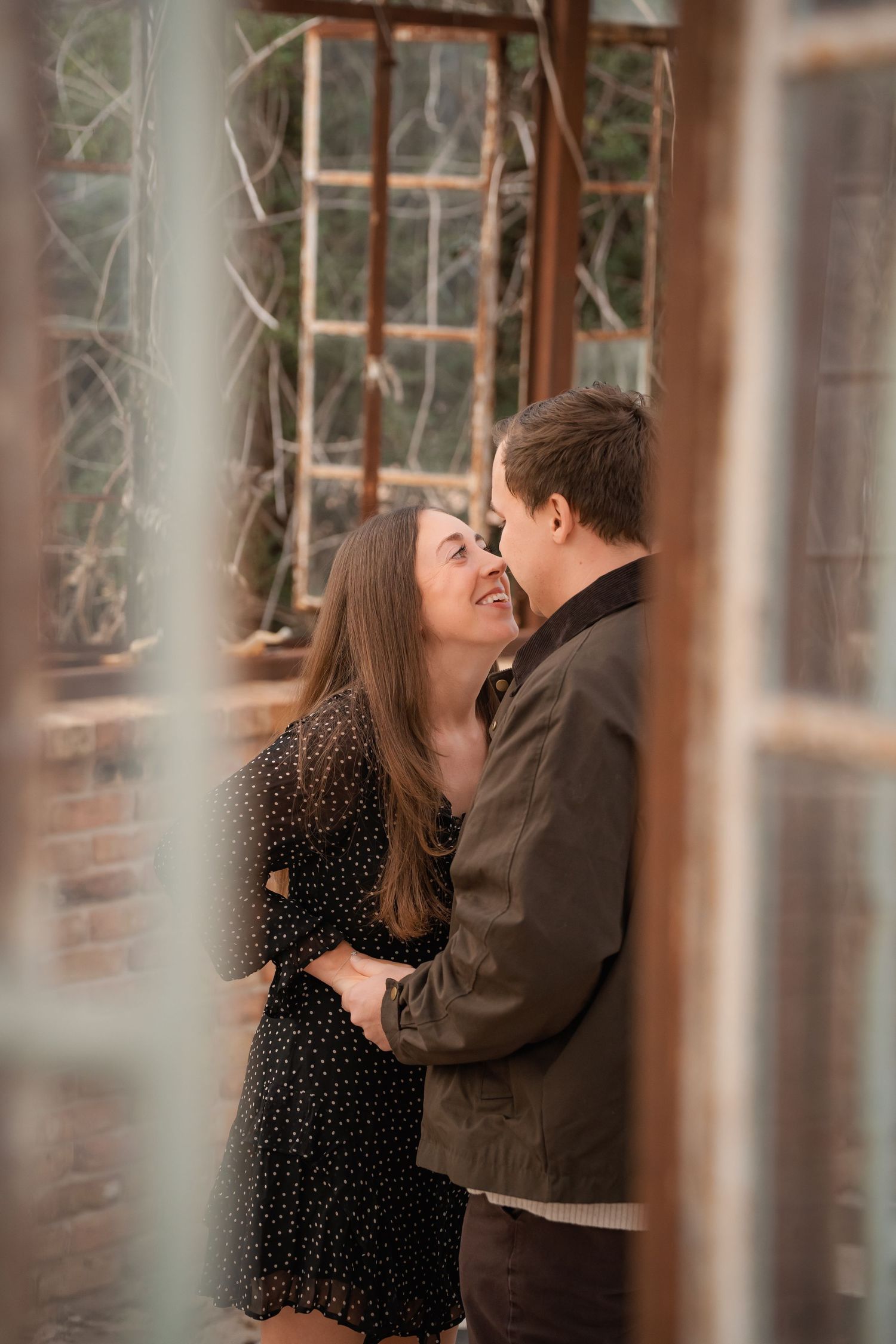 A couple shares a romantic moment, smiling at each other in an embrace near rustic windows.