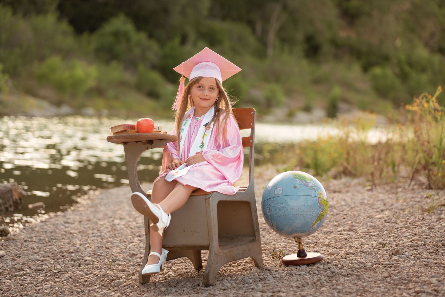 Young girl in pink graduation cap and gown sits on school desk beside globe outdoors near water.