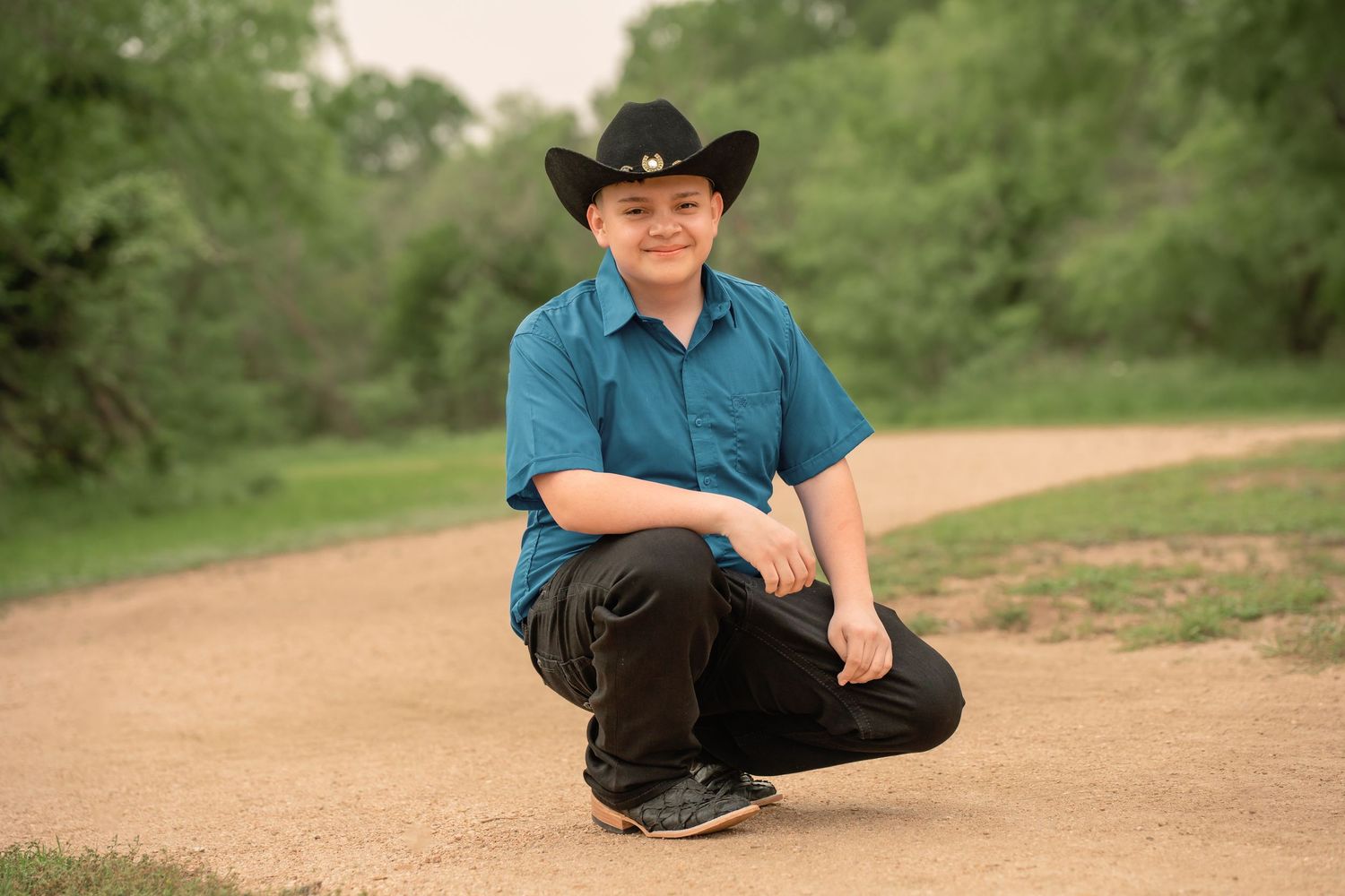 A young boy in a black cowboy hat and teal shirt crouches on a dirt path surrounded by green trees.
