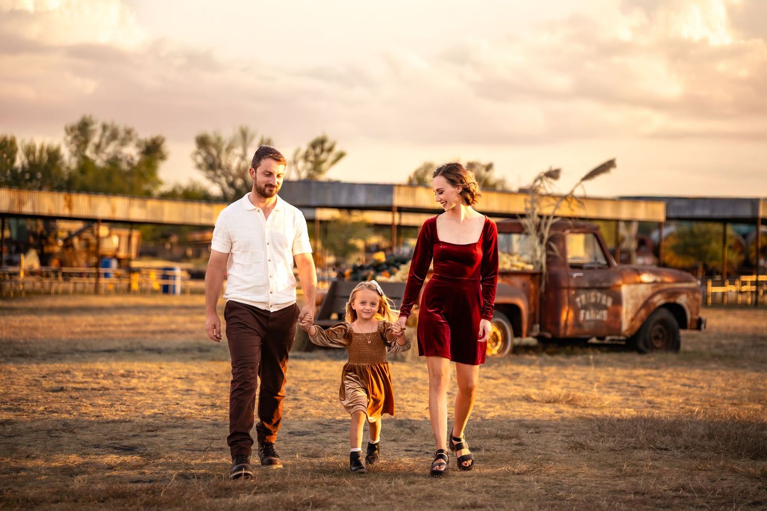 Family of three walking hand-in-hand at golden hour near a rustic farm with a vintage truck in the background.