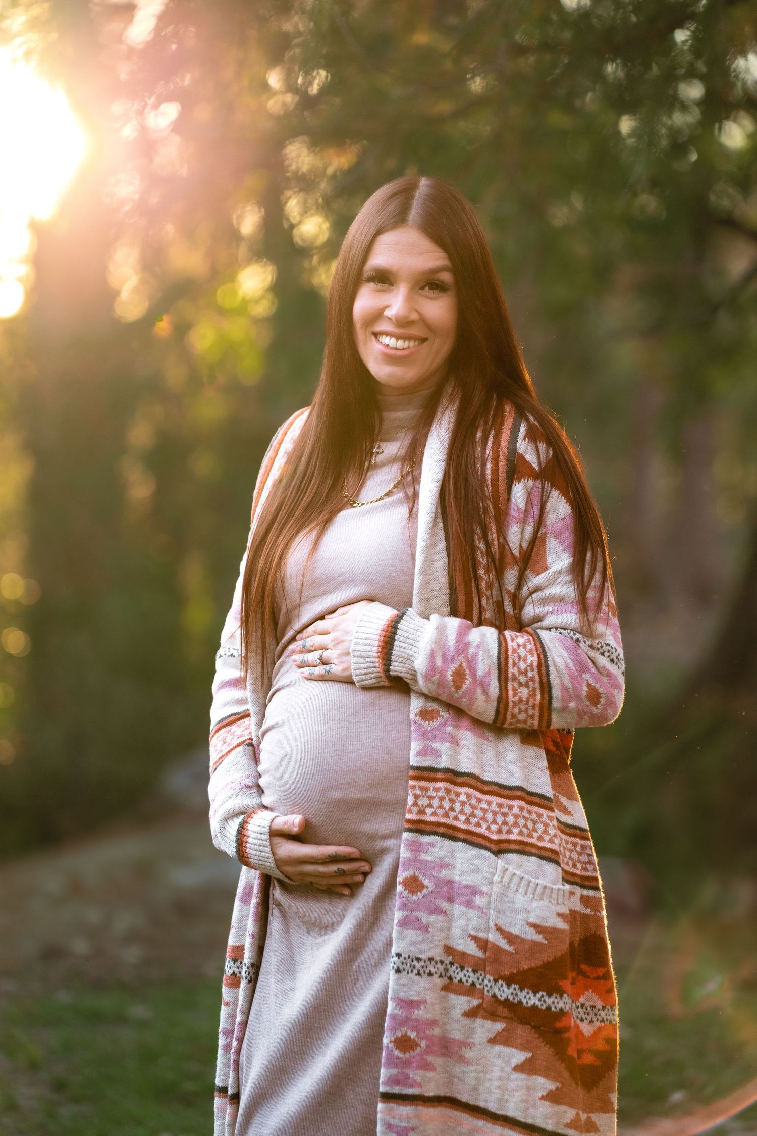 Smiling pregnant woman in boho cardigan cradling her baby bump outdoors in golden sunset light.