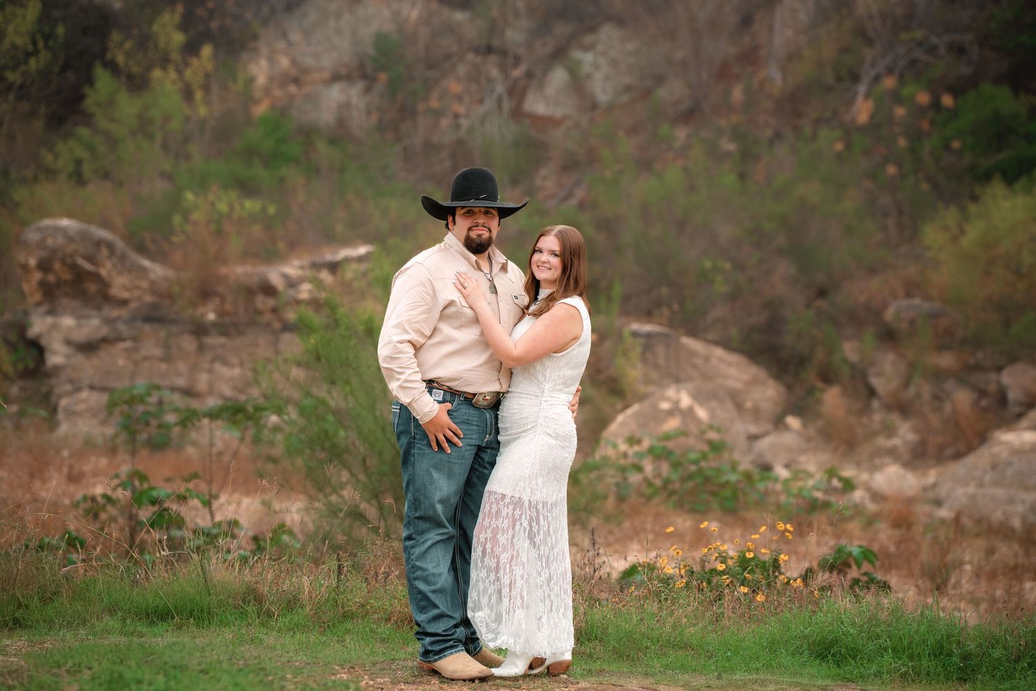 Couple posing outdoors in rustic setting, man in cowboy hat and woman in white lace dress, rocky landscape background.