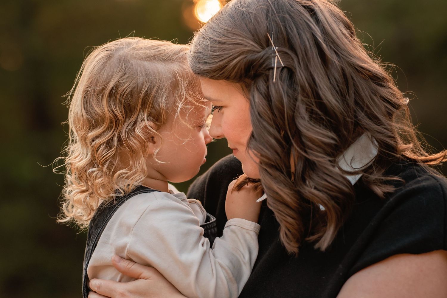 A mother and young child share a tender forehead-to-forehead moment outdoors in warm golden sunlight.