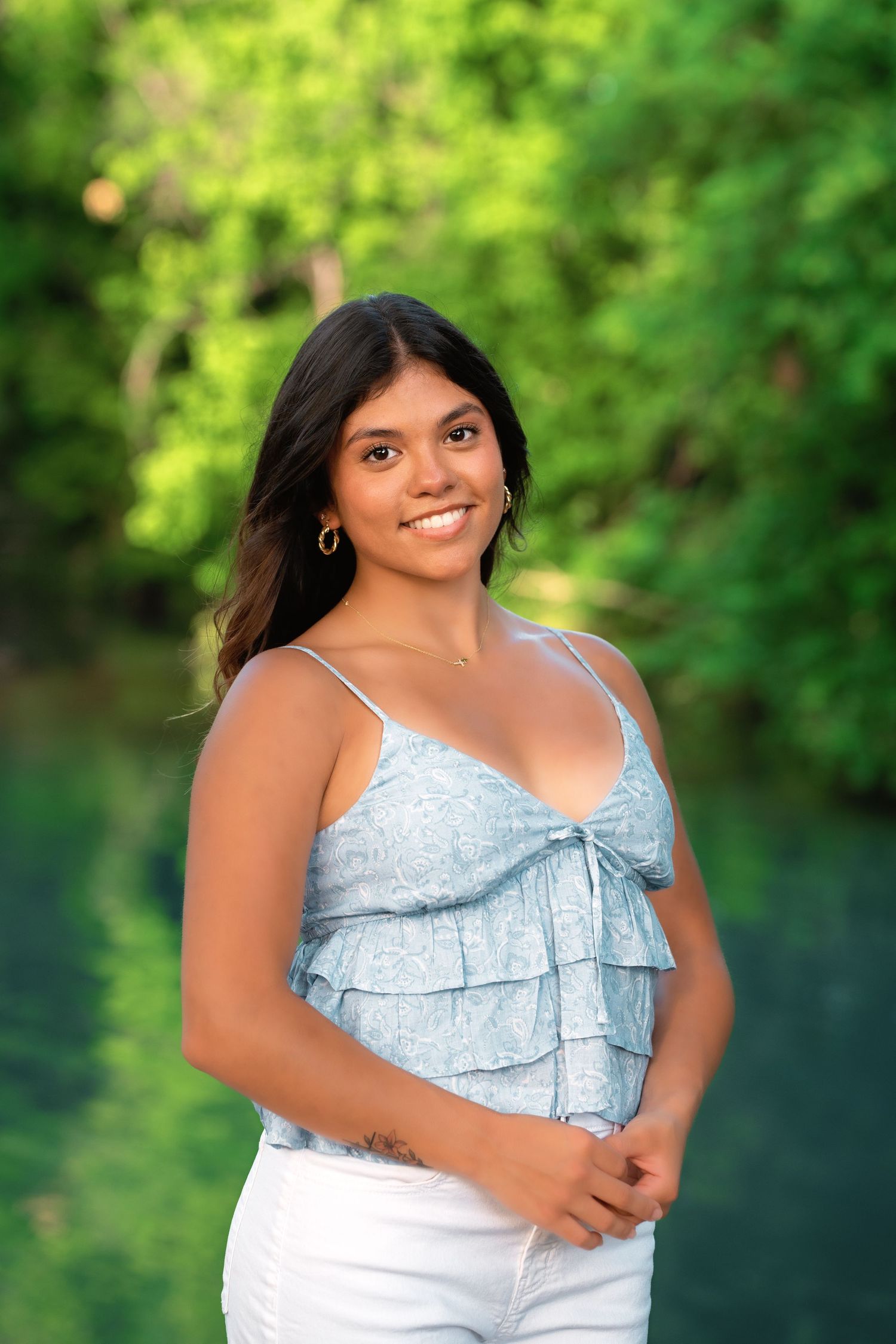 Smiling young woman in blue ruffled top and white pants posing outdoors near water with green trees.