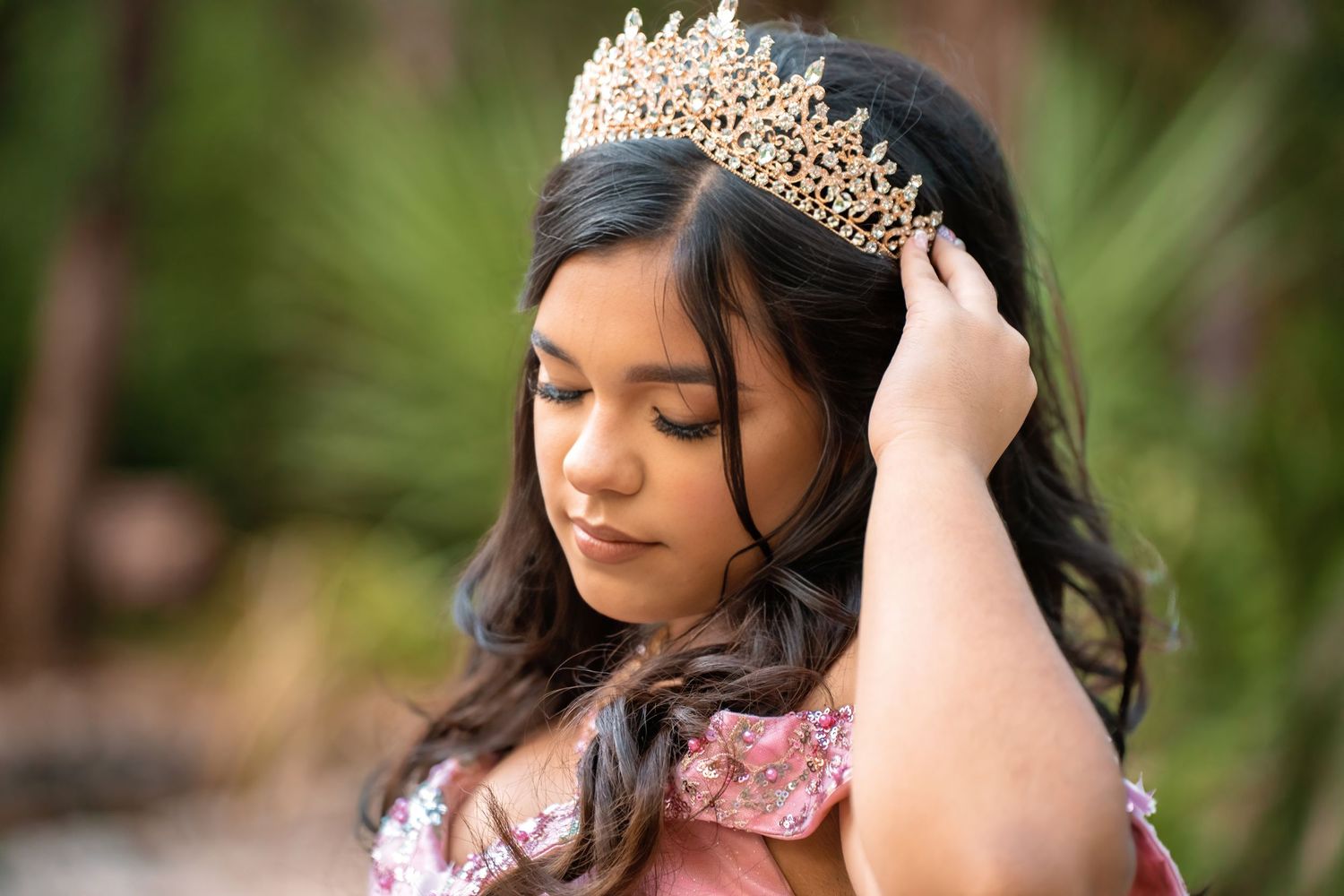 Young Latina girl adjusting gold tiara crown in her dark hair wearing a pink quinceañera dress outdoors.