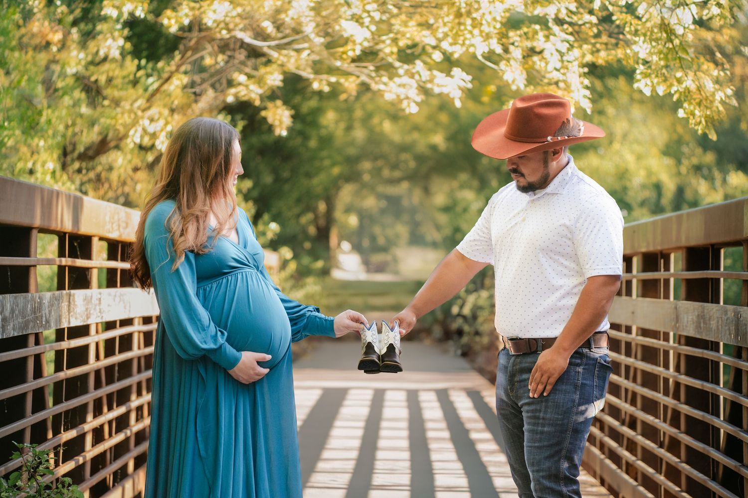 Pregnant woman in teal dress with partner holding baby shoes on a wooden bridge outdoors.