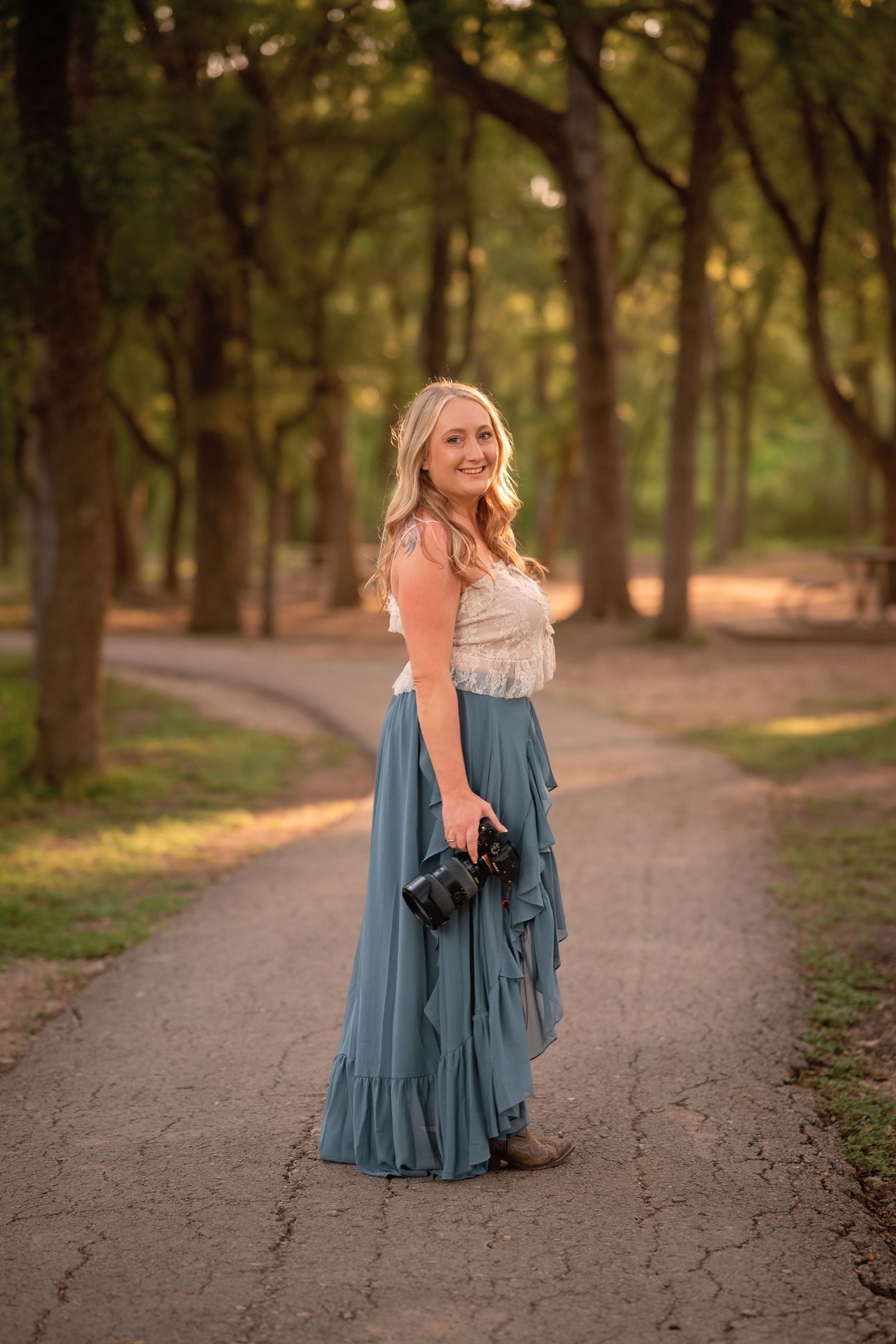 Blonde woman in teal skirt and lace top holds camera on tree-lined path in golden hour light.