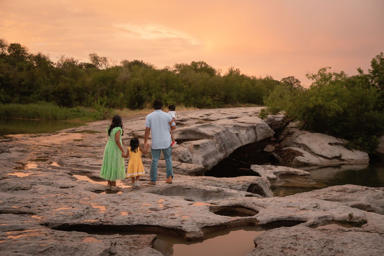 Family of three standing on rocky terrain watching a vibrant sunset over a serene river landscape.