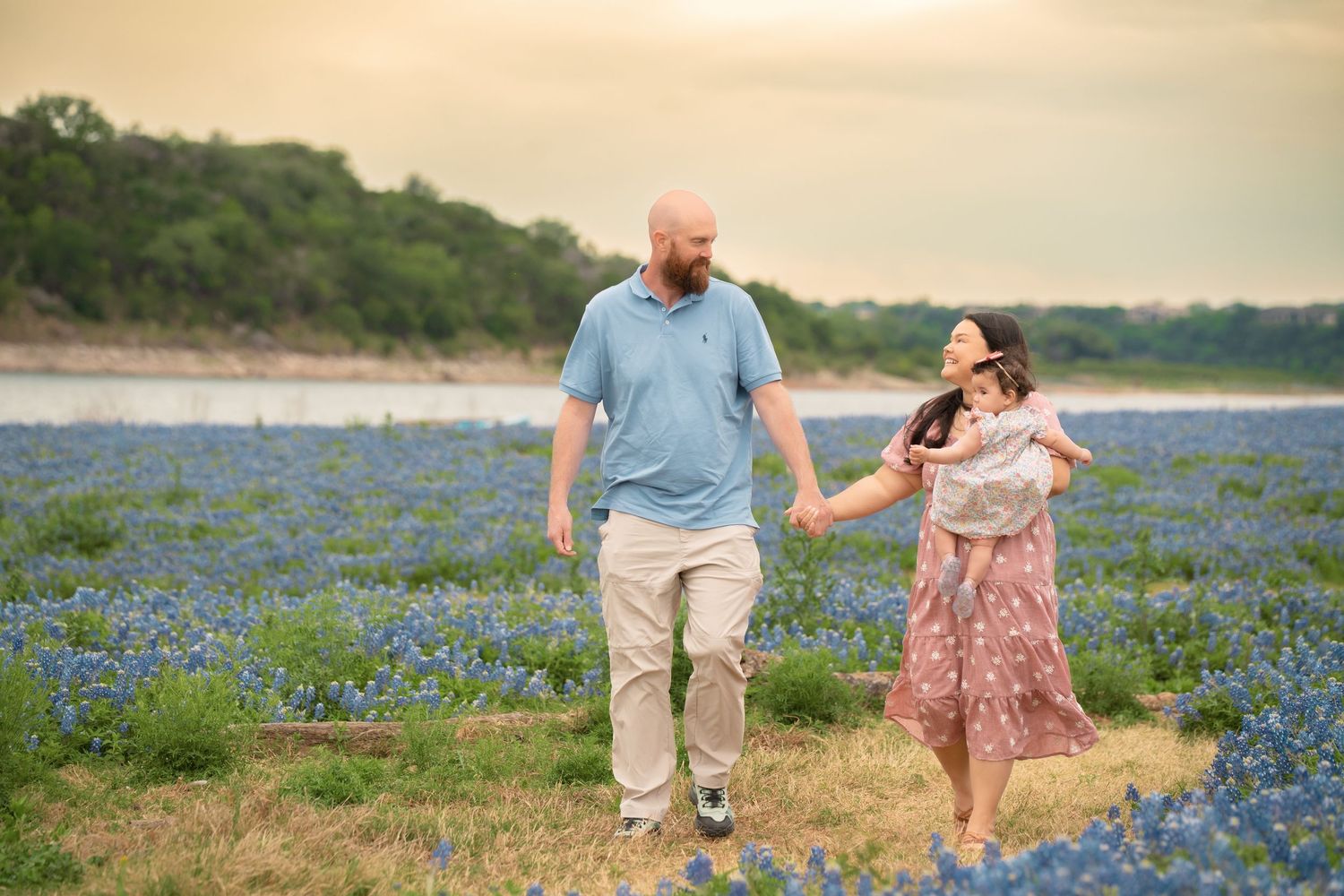 Family walking through a field of bluebonnets at sunset, holding hands with baby in Texas wildflower meadow.