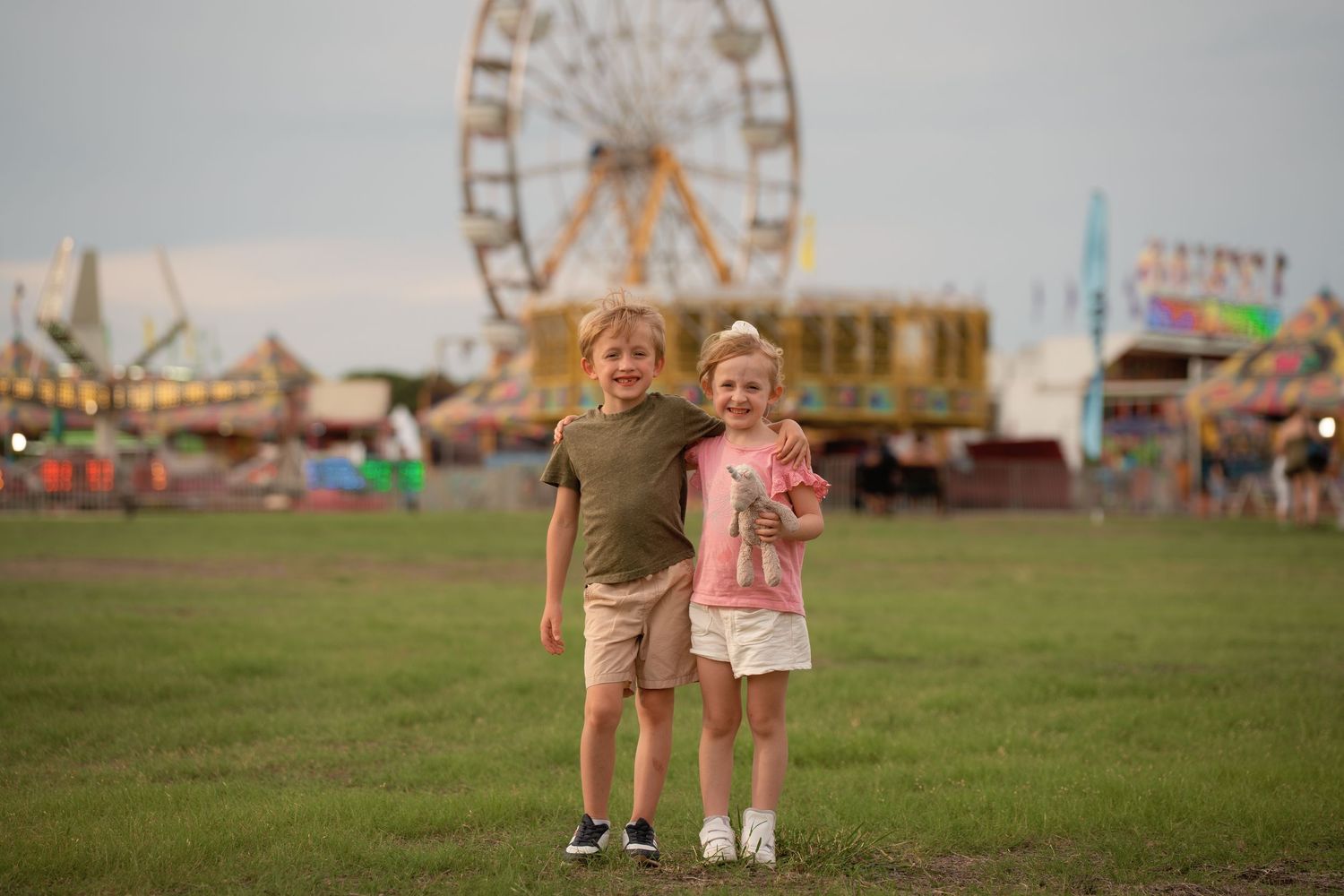Two children smile together at a colorful county fair with a Ferris wheel in the background at dusk.
