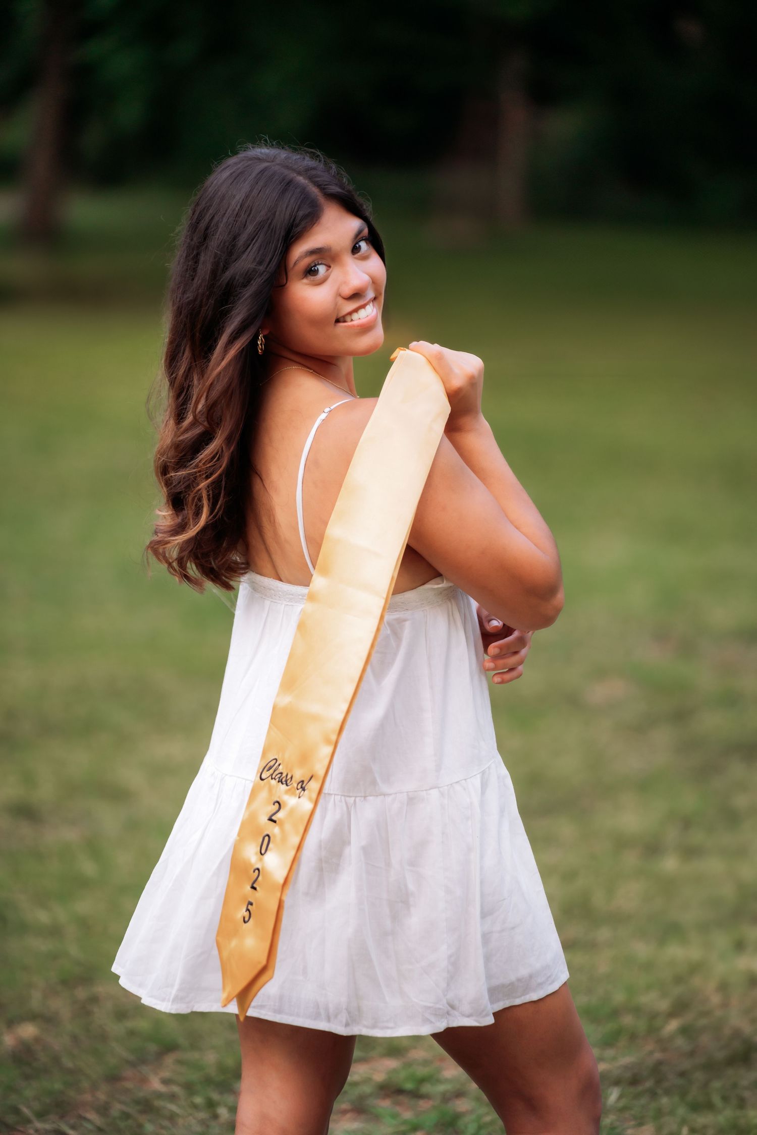 Smiling young woman in white dress wearing a gold Class of 2023 graduation sash, posing outdoors in a green park.