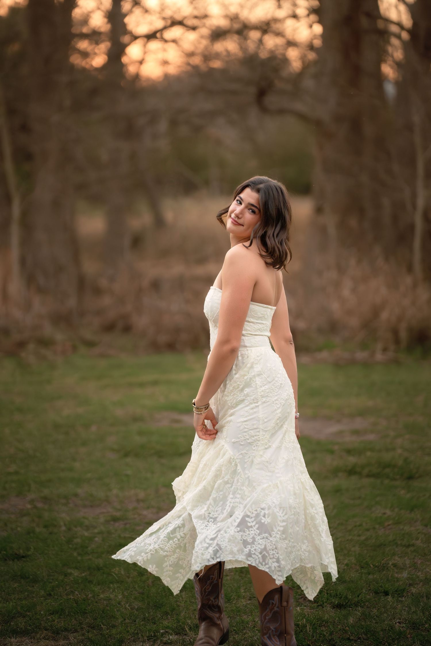 Woman in white lace strapless dress and cowboy boots twirling in a field at golden hour sunset.
