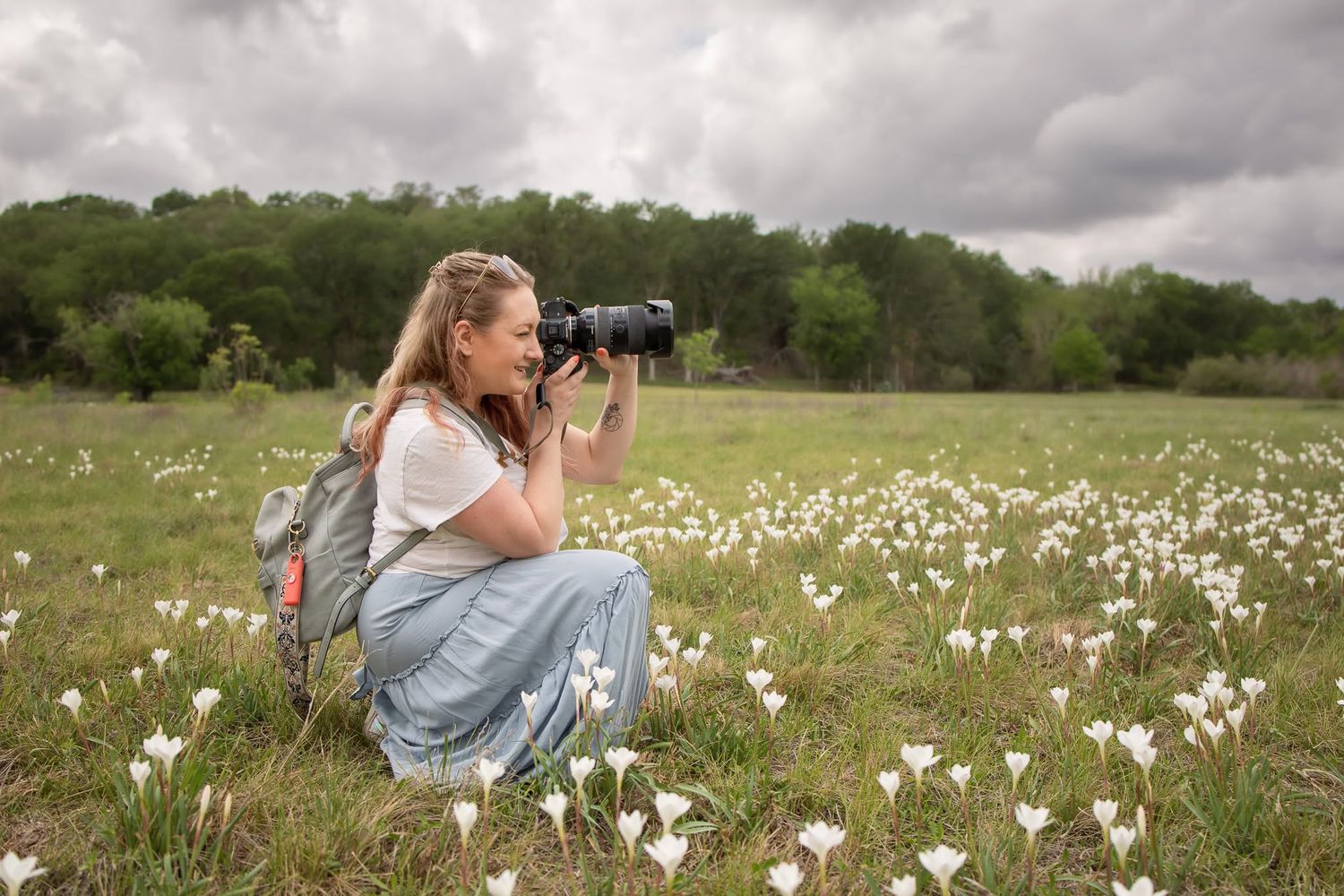 Woman photographer kneeling in a field of white wildflowers, capturing nature photos with a DSLR camera on a cloudy day.