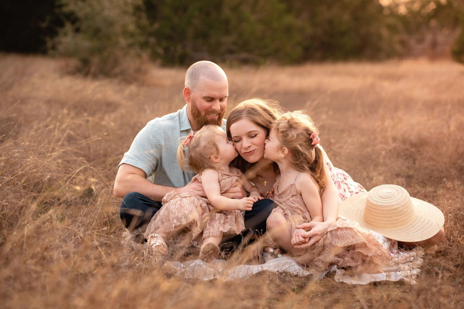 Family of four sitting in golden field, parents and two young girls in matching dresses sharing a tender embrace.