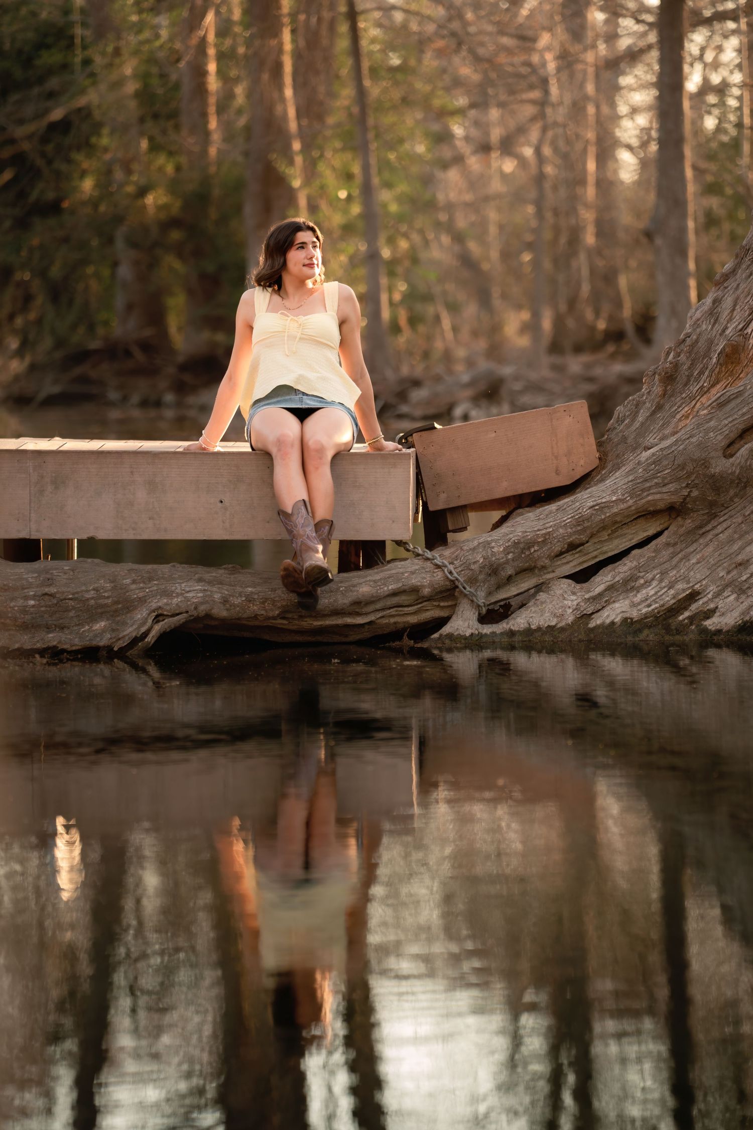 Young woman in cowboy boots sits on a wooden dock over a calm reflective lake surrounded by forest trees.