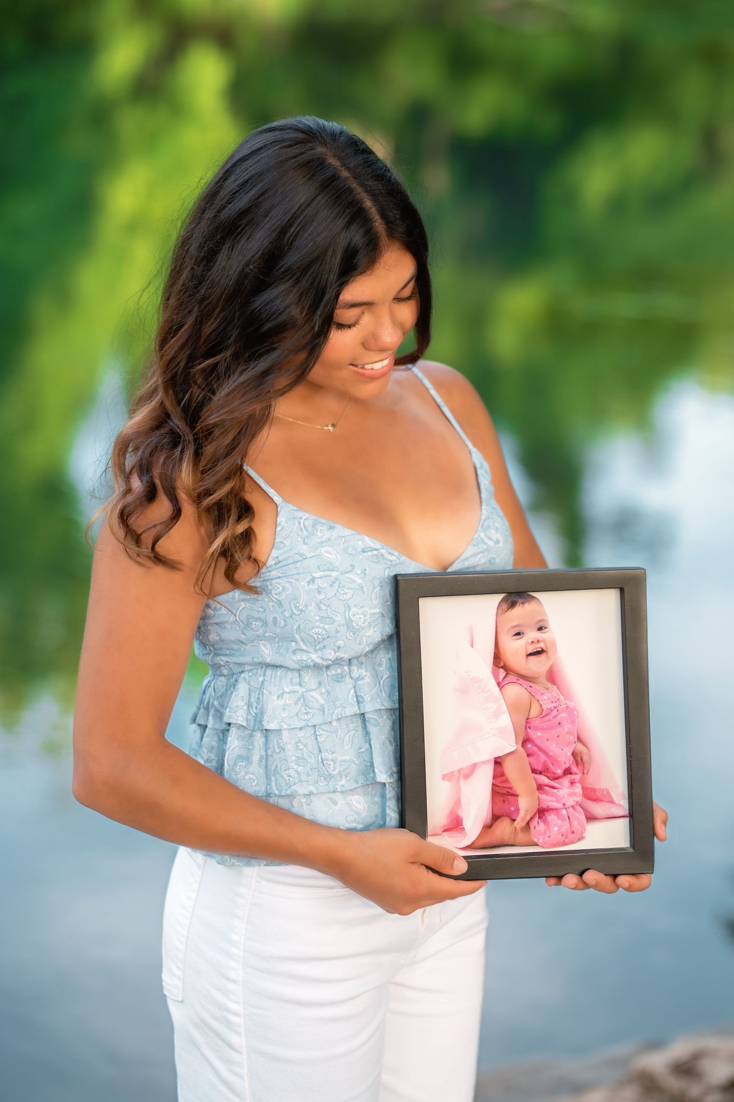 Young woman in blue top and white jeans smiles while holding a framed photo of a baby outdoors near water.