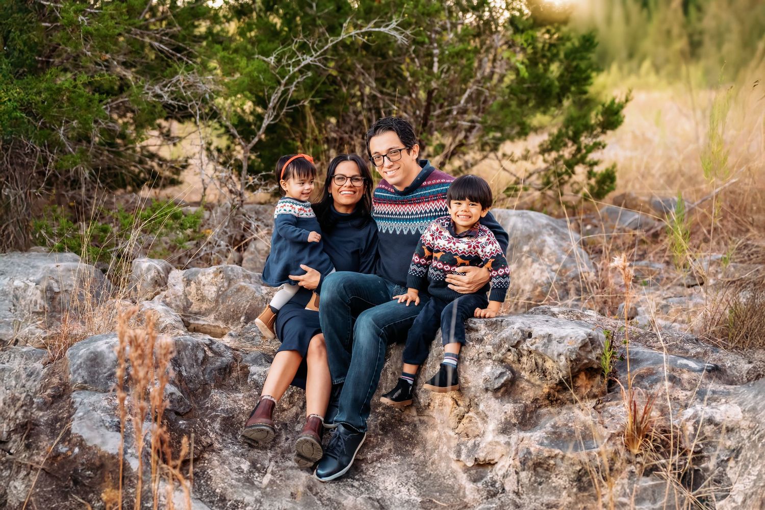 Family of four smiling together on rocks outdoors, surrounded by trees and dry grass in a natural setting.
