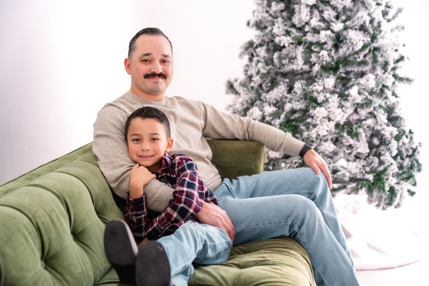 Couple cuddling and smiling on a green couch with a decorated Christmas tree in the background.
